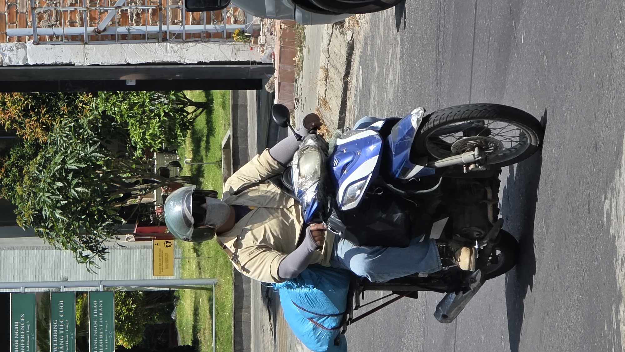 A man wearing a helmet, light brown jacket, blue jeans, and grey arm sleeves is riding a blue and white motorbike. He is traveling on a gray asphalt road. Strapped to the back of the motorbike is a large blue bag. To the left, a sign displays Vietnamese script and a partial view of foliage. The location is identified as Bắc Nha Trang, Vietnam.