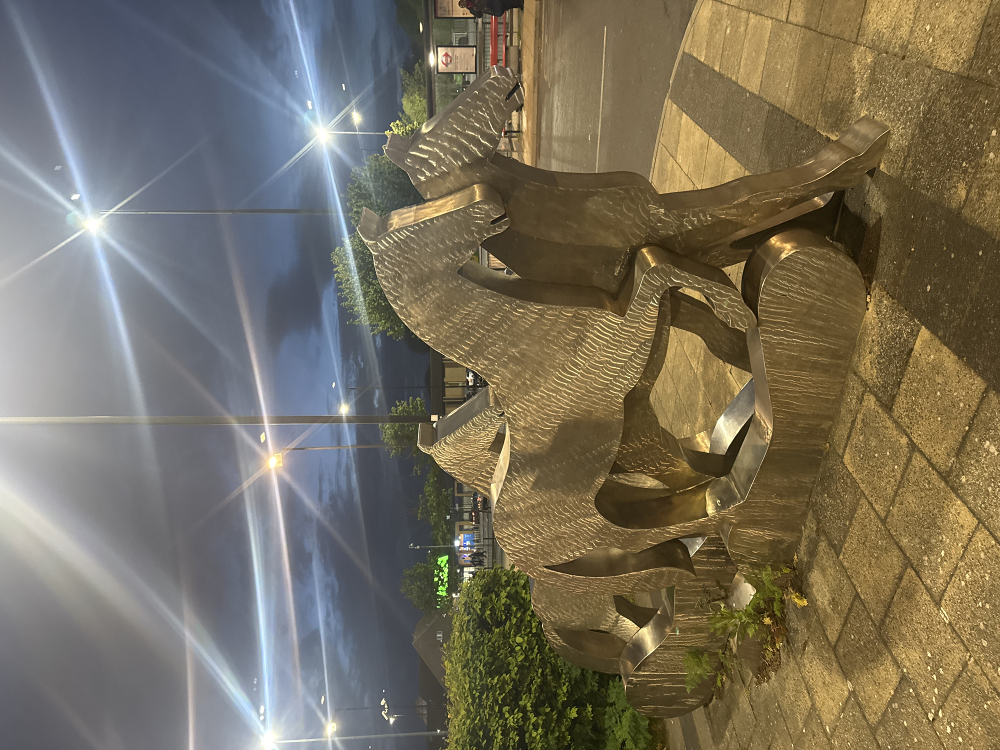The image displays an urban street scene at night in London, United Kingdom. In the foreground and midground, a prominent, large metallic sculpture captures attention. It appears to be made of brushed bronze or a similar reflective material, featuring a distinct textured, wavy surface. The sculpture's abstract, intertwined forms suggest stylized animals, possibly two horses or mythical creatures in dynamic motion, with flowing lines and shapes.
The sculpture rests on a paved area made of light-colored rectangular blocks, intersected by darker patterned strips. Green bushes and shrubbery are planted around its base, and small weeds can be seen growing from cracks in the paving. The dark blue-grey night sky is lit by several bright streetlights, which produce prominent starburst lens flares that cast light across the scene.
In the background, an asphalt road stretches out, with a building or structure visible beyond. This background structure features a recognizable red circular logo of the London Underground, serving as a clear location cue for London. A single person is partially visible standing near a red barrier in this distant area. Further into the background, a faint green glowing sign can be discerned, though its text is illegible. The overall impression is one of a quiet urban night, well-lit by artificial sources, highlighting the artwork in its public setting. The weather appears clear, despite the dark sky.