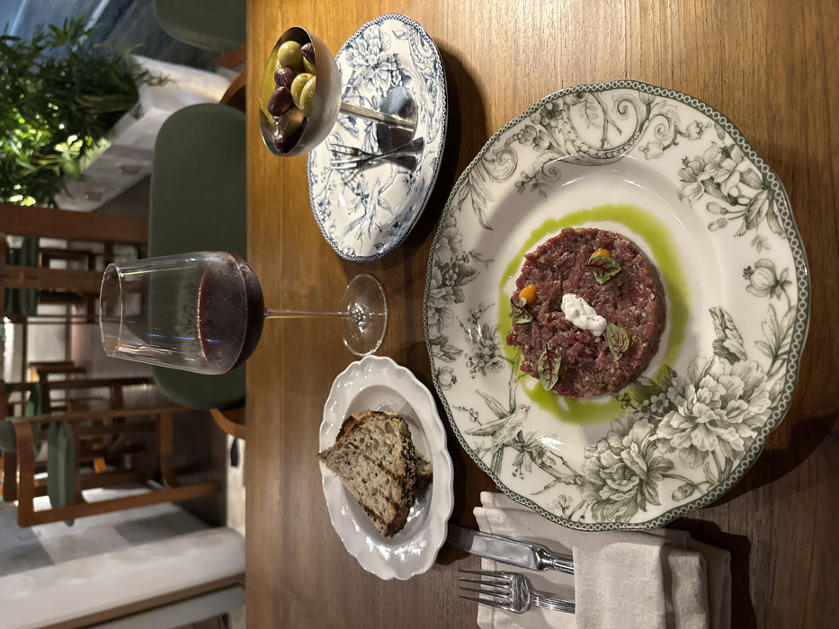 A dining table in a restaurant in Tibubeneng, Indonesia, is set for a meal. The wooden table displays a main course of steak tartare, presented on a white plate with a green and white floral and bird pattern. The tartare is garnished with a white dollop, small orange elements, and red-veined microgreens, with a green sauce encircling it. To the left, a small white scalloped plate holds a slice of dark, rustic bread. To the right, a stemmed wine glass contains red wine. Further right, a small silver bowl on a pedestal holds green and black olives, resting on an oval blue and white patterned plate with a fork and knife beneath it. A set of cutlery, a fork and knife, are wrapped in a plain white napkin to the left of the main dish. In the background, out of focus, are wooden high-backed chairs with green upholstery and a planter with green foliage, indicating an indoor dining environment. No individuals are present in the frame.