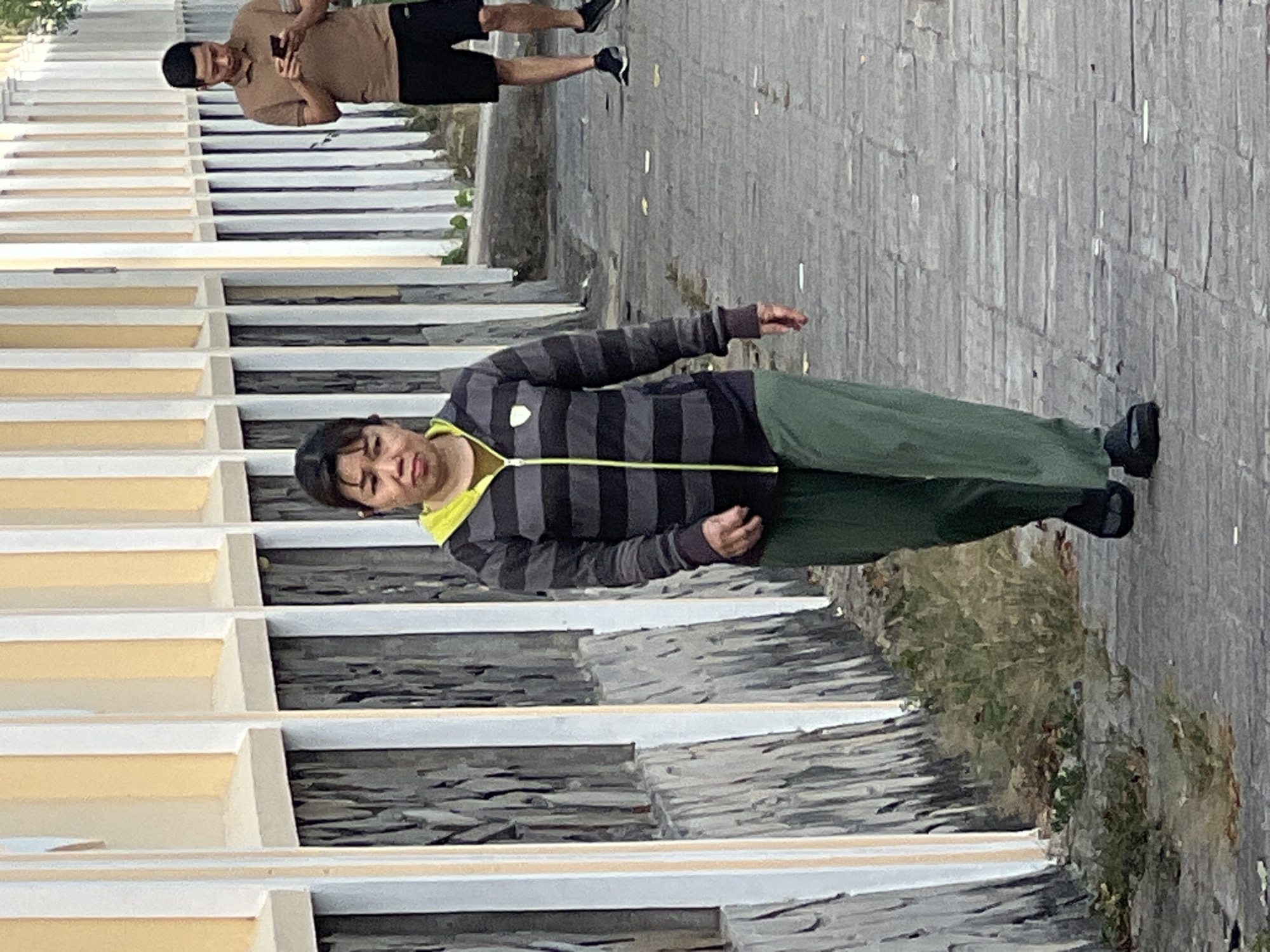 An outdoor, eye-level, medium shot captures two individuals walking on a paved walkway beside a wall. The wall, on the left, is constructed with alternating vertical bands of yellow and white pillars, with a textured gray surface beneath. To the right, a man in a brown polo shirt and black shorts walks away from the camera, looking at his phone. In the foreground, a woman with dark hair and bangs walks towards the camera, wearing a horizontally striped zip-up hoodie with gray and black stripes and a bright lime green zipper and lining, and loose-fitting green pants. She is wearing black sandals. The ground is paved with gray rectangular stones and littered with some fallen leaves. Vegetation, including dry grass and bushes, grows alongside the wall. The lighting suggests it is daytime. The scene appears to be a public walkway in Đà Nẵng, Vietnam, possibly near a coastal area due to the open sky and walkway.