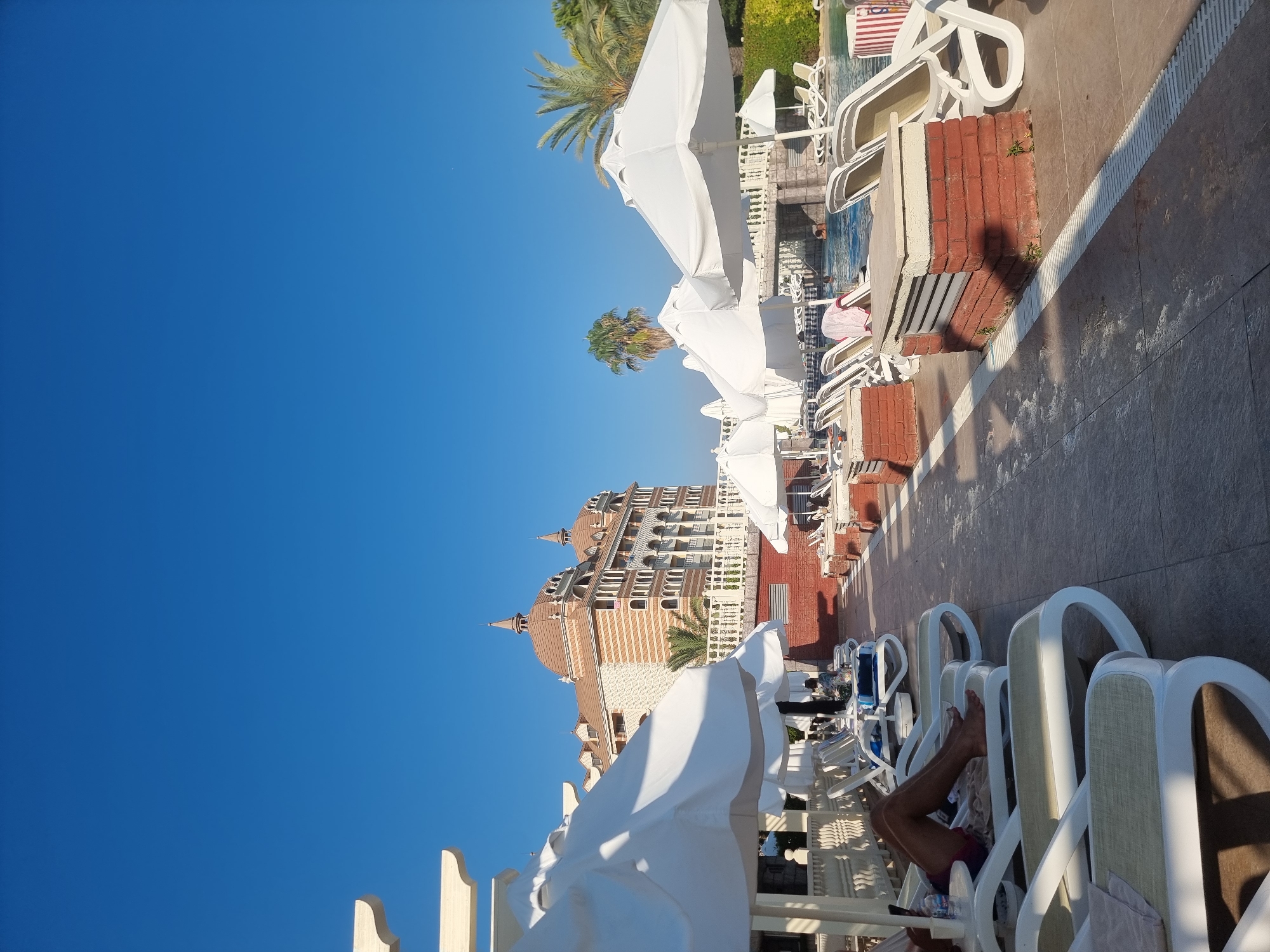 The image depicts an outdoor swimming pool area at a resort in Manavgat, Turkey, on a clear, sunny day. The foreground features rows of empty white lounge chairs with light-colored cushions, some with towels draped over them. White umbrellas are erected over some of the lounge chairs, providing shade. A person's legs and torso are visible, reclining on a lounge chair on the left side of the image, holding a mobile phone.
In the middle ground, the swimming pool is visible with some people swimming. A decorative bridge crosses over a section of the pool. Palm trees and lush green vegetation are present behind the pool area.
The background is dominated by a large, ornate hotel building with distinct architectural features, including multiple arched windows, decorative columns, and domed roofs. The hotel is designed with a patterned facade featuring horizontal stripes. White balustrades and brick structures are also visible around the pool and hotel. The sky is a clear, vibrant blue. The overall scene suggests a tranquil resort atmosphere.
