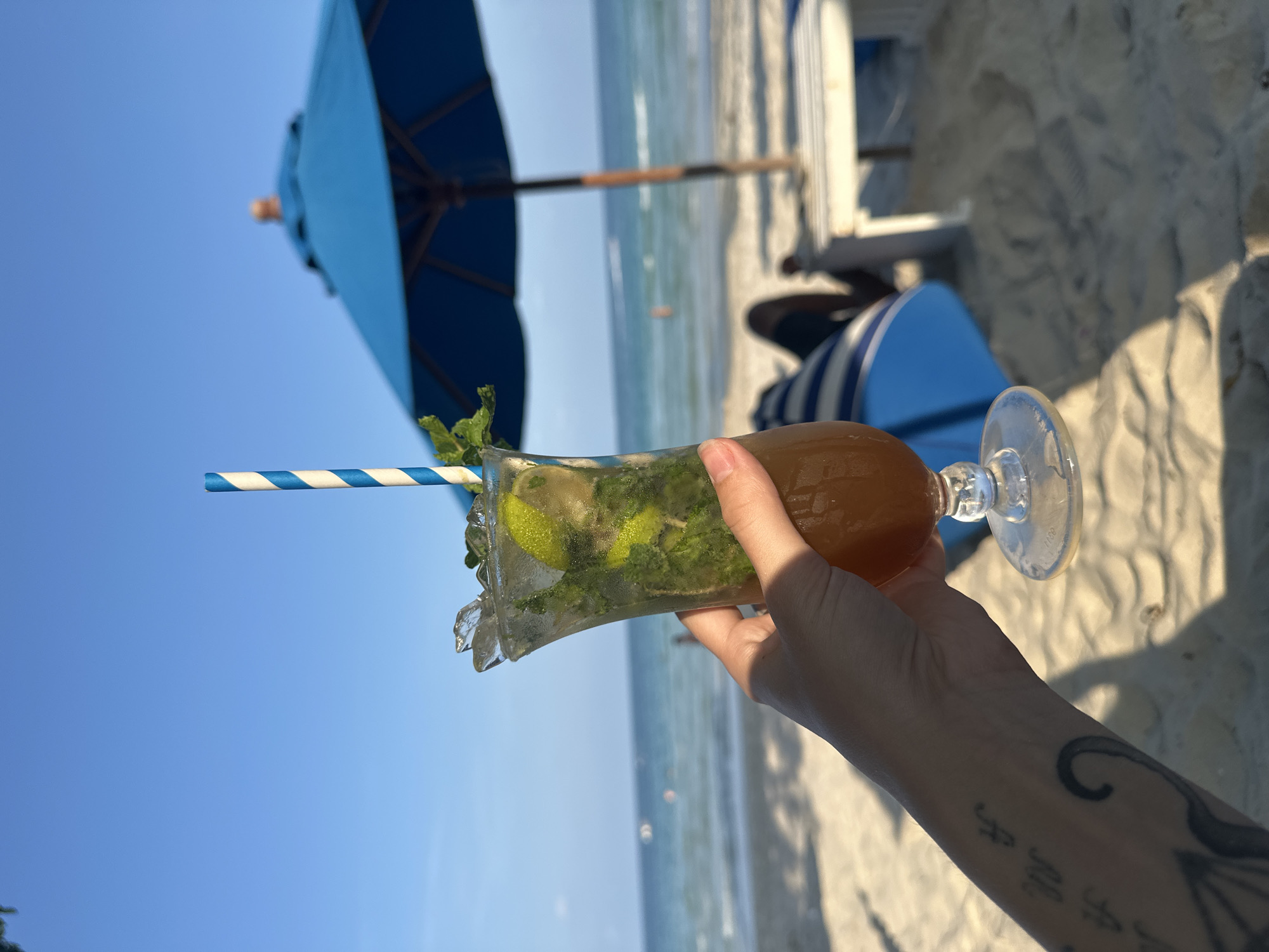 A person's tattooed arm extends upward, holding a tall glass filled with a mojito, complete with a blue and white striped straw and mint leaves. The drink is set against the backdrop of a beach in Baan Chaweng, Thailand. A bright blue sky is visible, as well as the ocean, beach sand, and an open blue umbrella in the background.