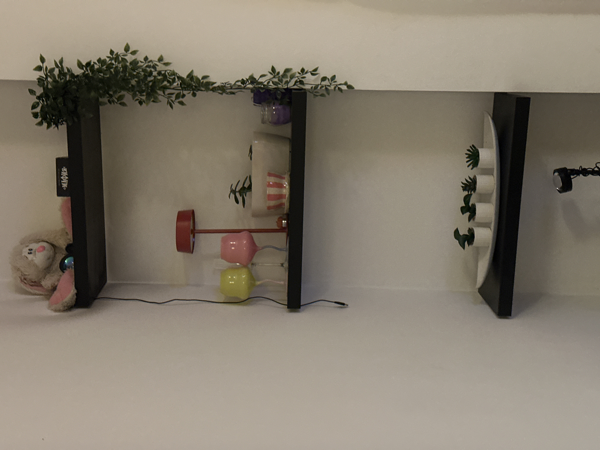 The image depicts a shelf arrangement against a white wall. From left to right: a stuffed toy bunny sits next to a black shelf upright with a small, dark label on the shelf. Above the shelf is greenery. Adjacent to this shelf, another black shelf holds an assortment of objects. There is a stack of potted plants, with purple, white, and pink planters. Below the plants, is a red and pink lamp with a yellow light bulb. Adjacent to this shelf, on the right, is a white plate containing small succulent plants. Next to the plate, is another black shelf. A flexible black cord is to the right of this shelf. The location of the image is not apparent, and cannot be linked to Novi Sad, Serbia.