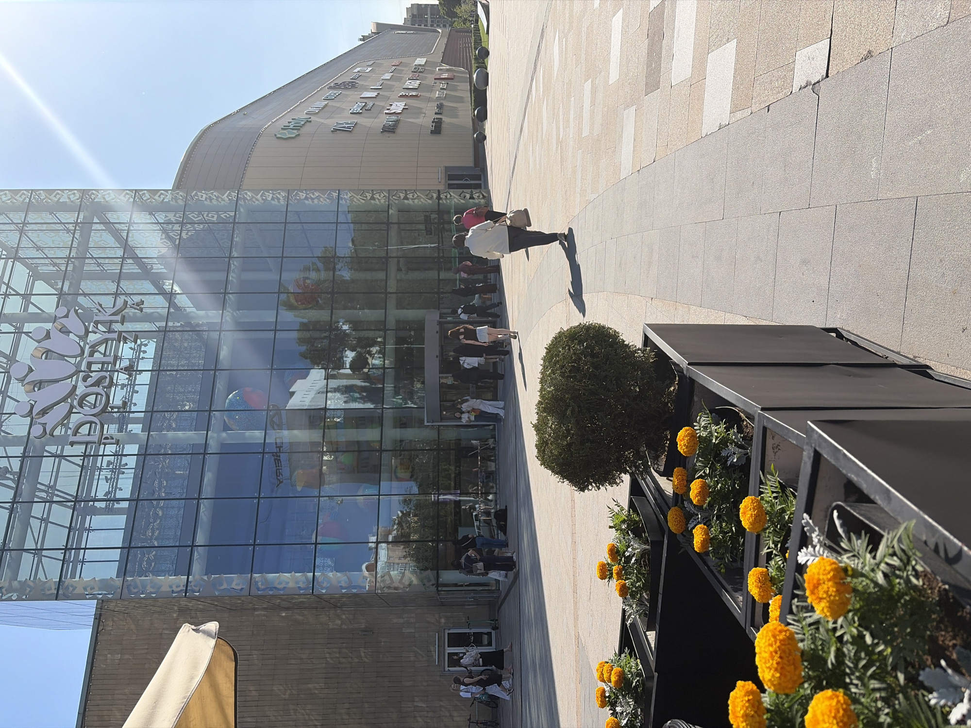 The image shows an exterior view of a building with a glass facade, presumably a commercial establishment or shopping mall, located in Almaty, Kazakhstan. A line of people are waiting to enter, suggesting potential access restrictions or interest in the facility. The scene is taken at ground level, adjacent to a pedestrian walkway paved with light-colored stone. Several planters containing vibrant yellow flowers line the walkway, enhancing the aesthetic appeal. The building's facade features multiple glass panels and reflective surfaces, mirroring the environment and reflecting blue from the sky. A secondary, beige-colored building is visible in the background. The lighting suggests it's daytime, and there may be sunlight or glare, given the presence of strong highlights in the frame.