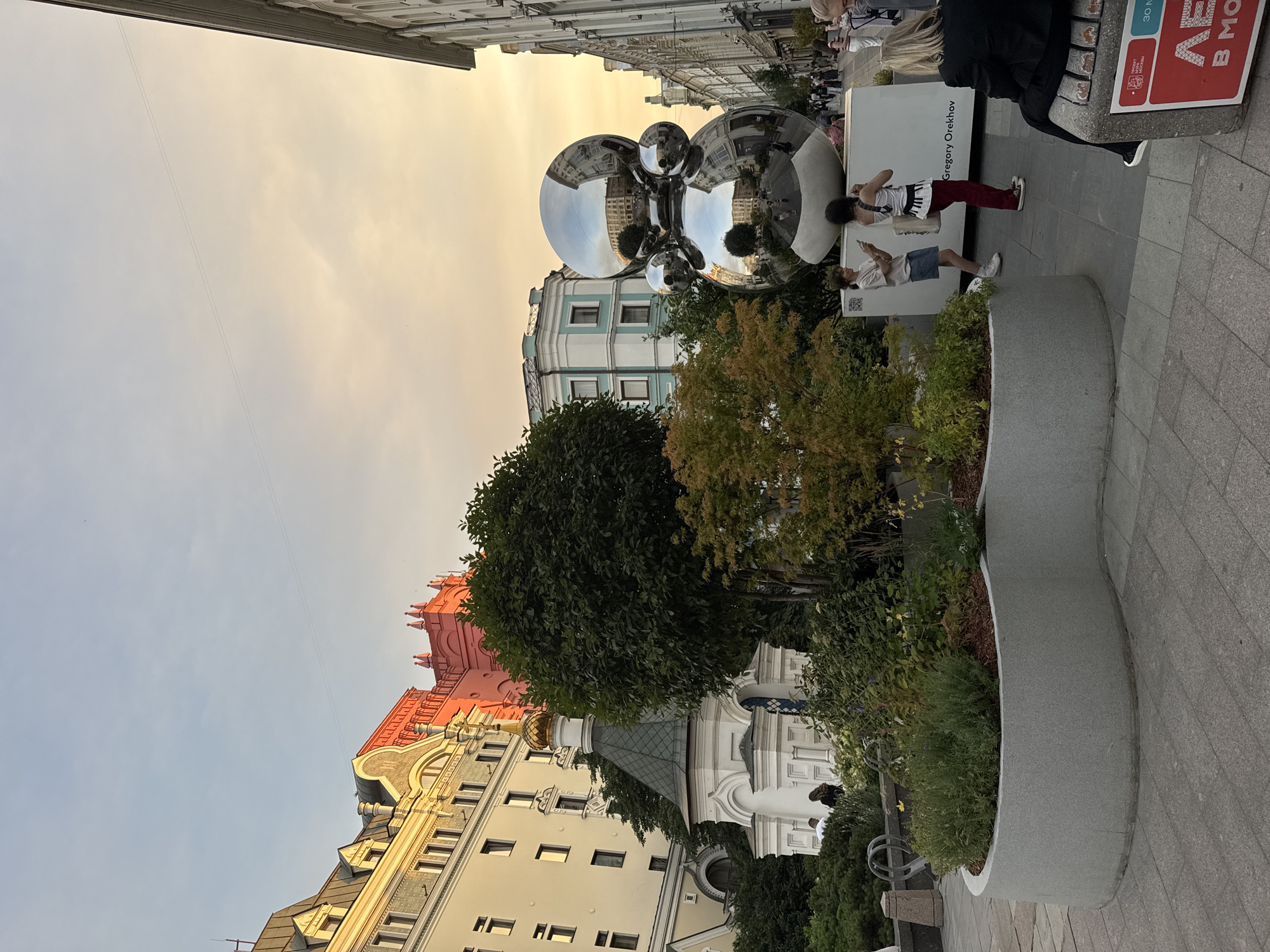 This image captures a street scene in Moscow, Russia, likely during the late afternoon or early evening, indicated by the warm, soft light bathing the sky and buildings. The focal point is a large, polished, spherical sculpture, reflecting the surrounding architecture and the sky.
To the left of the sculpture, a small white building with a golden dome, resembling a chapel or a small church, is visible. It is surrounded by lush greenery, including trees and bushes. Behind the chapel and to the left, a multi-story building with a light-colored facade and dark-tiled roofs stands prominently. Further to the left, a striking pink building with ornate architectural details rises, its color contrasting with the more subdued tones of its neighbors.
In the foreground, a curved, grey planter box filled with various plants and mulch is present. Two young women are standing near the sculpture, one with dark hair wearing a white top and dark pants, and the other with lighter hair wearing a light-colored top and a denim skirt. They appear to be interacting with their phones. To the right of the sculpture, a street lined with buildings continues into the distance, with several people walking along the sidewalk. A bench with a person seated on it is visible in the lower right corner, featuring a red sign with Cyrillic lettering. The overall impression is a blend of historic architecture, modern art, and urban life.