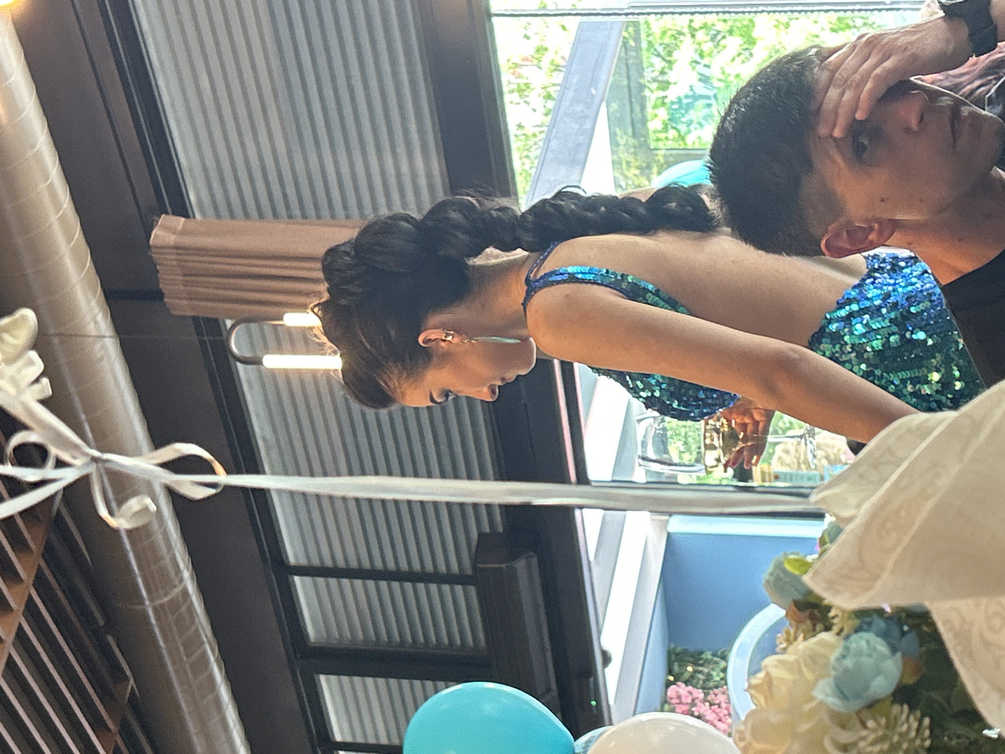 The image shows a scene indoors, possibly at an event or gathering. Two individuals are visible: a woman with dark hair braided and wearing a sparkling blue dress, and a man standing behind her. The man is touching his head. The background includes a window with a view of greenery, suggesting an outdoor location. There are balloons and floral arrangements in the foreground. The interior setting appears to have metal structures and a modern design. The location in Burgas, Bulgaria, cannot be determined from the image.