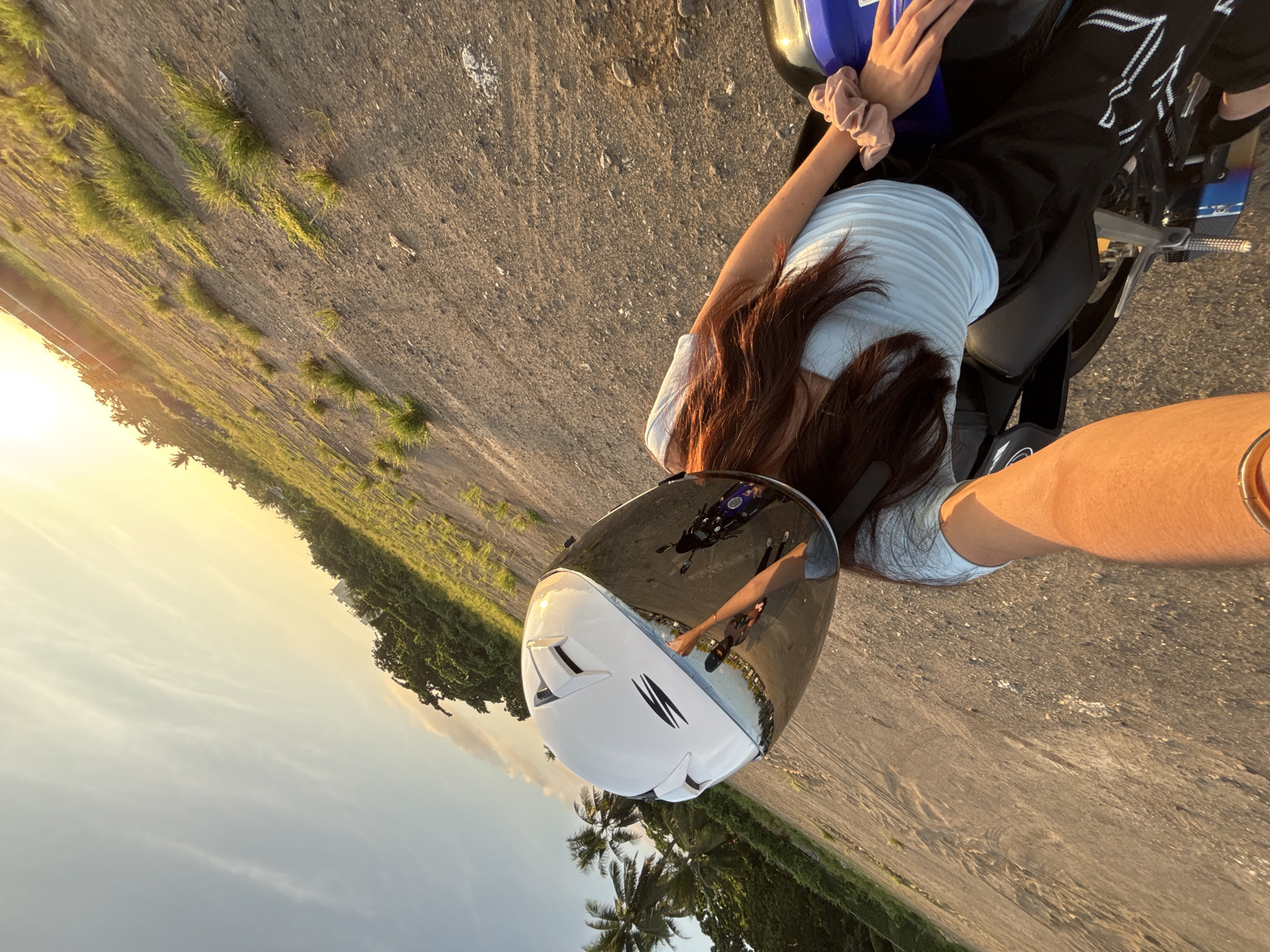 A person wearing a white motorcycle helmet with a reflective visor is seated on a motorcycle. The helmet's visor reflects a partial image of the rider, the motorcycle, and the surrounding environment. The rider has long, dark brown hair and is wearing a light blue ribbed top and black pants with white accents. A pink scrunchie is visible on their wrist. The motorcycle is blue and black.
The scene appears to be an open, unpaved area, possibly a dirt field or undeveloped land, with sparse vegetation and dry grass growing in patches. The ground is a mix of dirt and small stones. In the background, there is a line of lush green trees, including palm trees, suggesting a tropical climate. Beyond the trees, a few buildings are visible, hinting at the edge of a populated area.
The sky is a gradient of soft blues and warm yellows, indicating either sunrise or sunset. The low angle of the sun casts long shadows and illuminates the scene with a warm, golden light. The overall impression is one of a casual ride or a pause during a motorcycle excursion in a somewhat rural or undeveloped part of Davao City, Philippines, given the presence of palm trees and the general landscape.