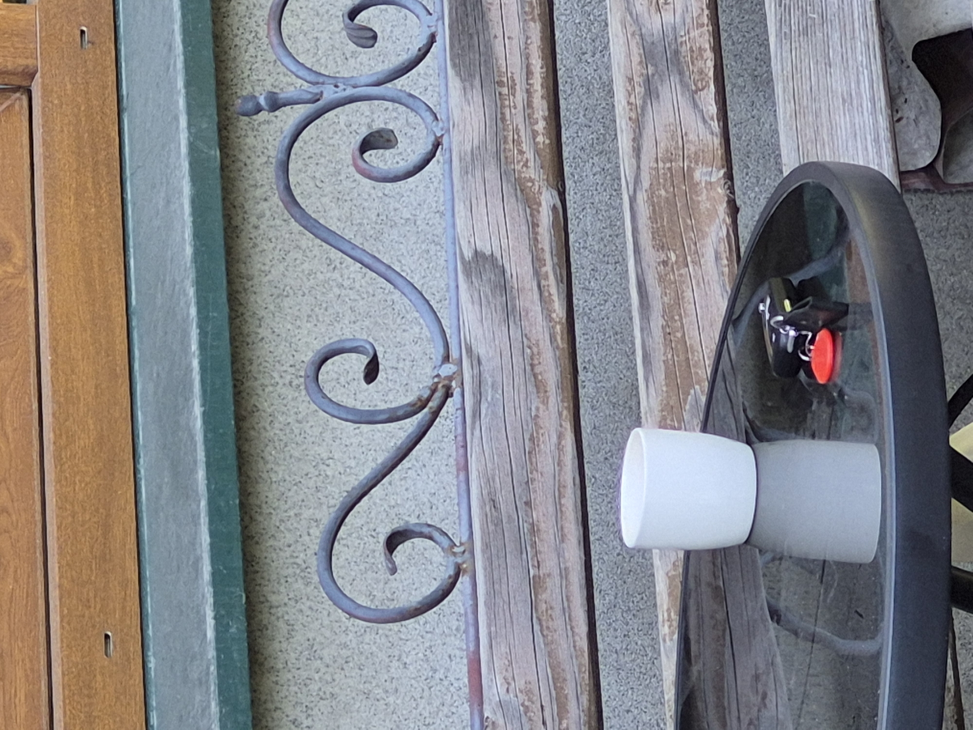 A white cylindrical cup sits on a round, dark, reflective table. Next to the cup, on the table, are a set of keys with a red fob. The table is positioned in front of a weathered wooden bench. Behind the bench is a decorative wrought-iron railing with scrolled designs. The railing is mounted on a textured gray wall. Above the railing is a wooden structure and a green band. The scene is outdoors.