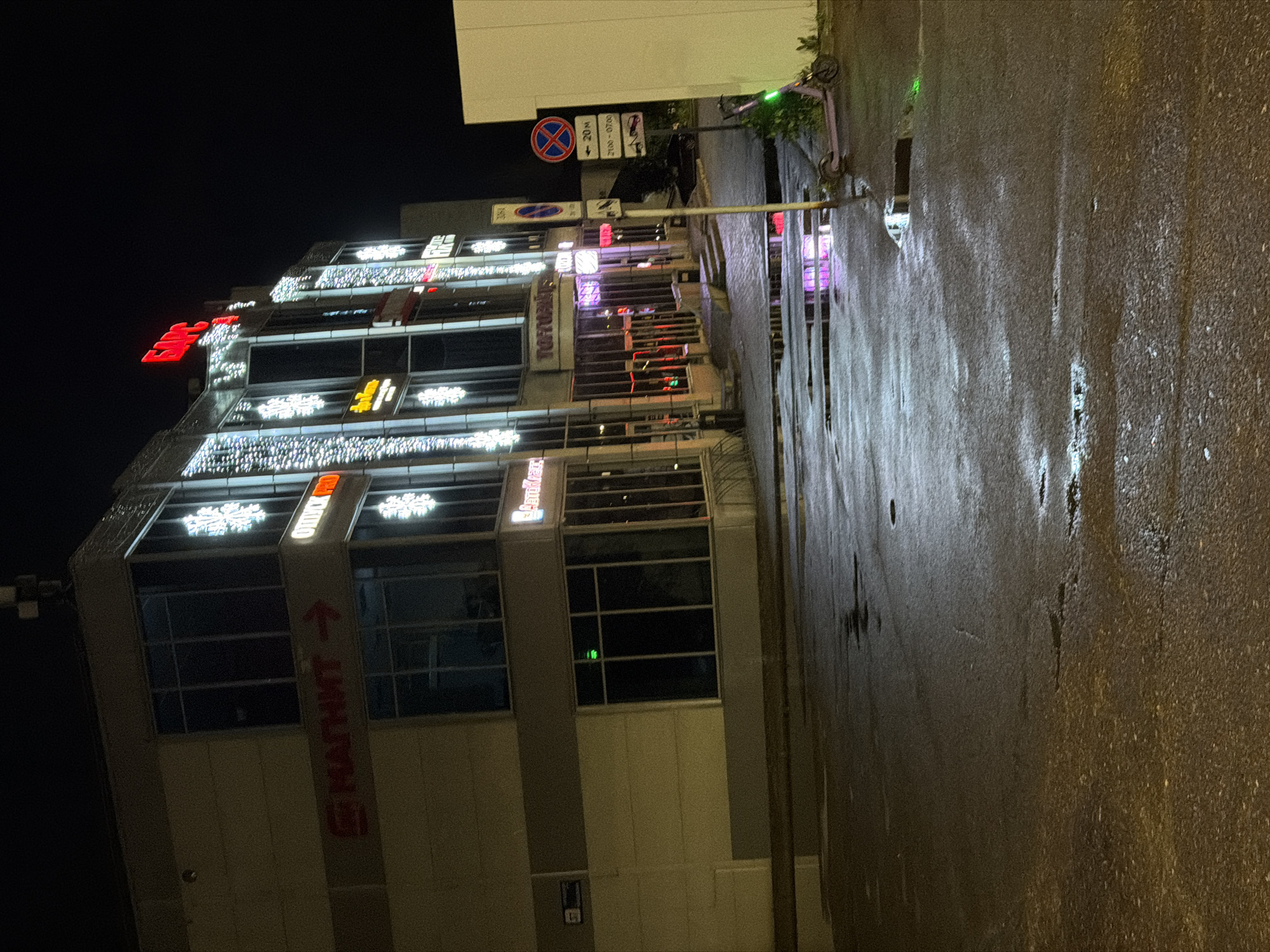 This is a nighttime, eye-level, medium shot of a modern building in Nizhnekamsk, Russia. The building, illuminated by festive lights and illuminated signage, features a facade with large windows and decorative snowflake motifs. Notable signs include "МАГНИТ" (Magnit, a Russian supermarket chain) with an arrow, and other businesses identified by neon lettering such as "БАРС" (Bars) and "ТОПТИАН" (Toptian).
The ground in front of the building is wet and reflects the lights from the building, indicating recent rain or puddles. A purple electric scooter with a green light is parked to the right of the frame, near a street signpost. The street signpost displays multiple signs, including a circular red and blue traffic sign with a white cross, an arrow pointing to the right with "20 M" below it, and time-based parking restrictions. The overall scene suggests a commercial or entertainment district within the city. There are no people visible in the image.