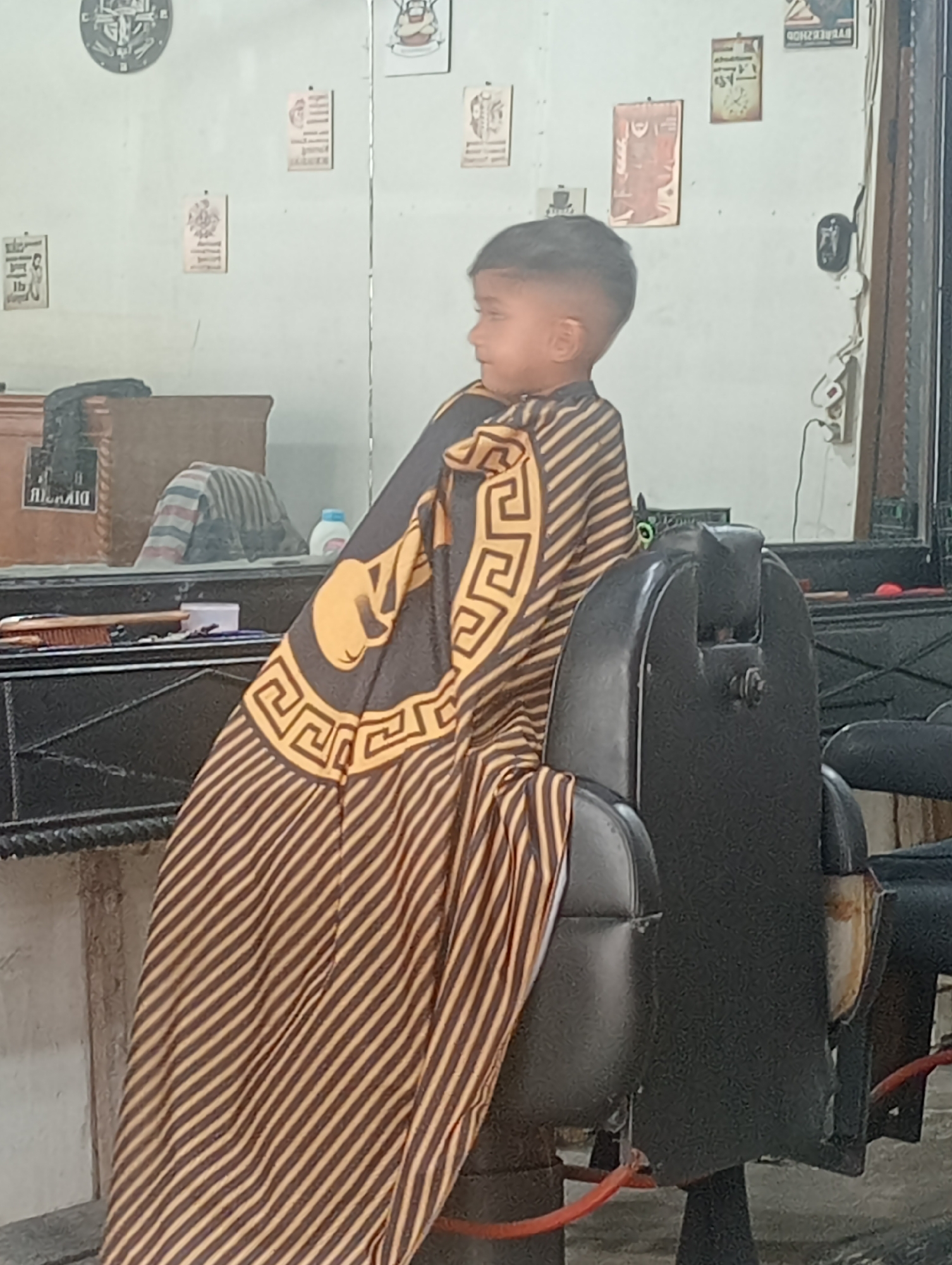 The image depicts a young boy in Pulorayeuk, Indonesia, sitting in a black barber's chair and draped with a cape featuring gold and black stripes along with a Greek key design and a gold face. The setting appears to be a barbershop, with a counter visible to the left of the chair, holding a brush and other grooming items. A mirror is in the background, reflecting the interior of the shop, including furniture and various decorative items or posters on the wall. A clock is also visible on the upper left of the wall. Electrical outlets with connected devices are on the right side of the image.