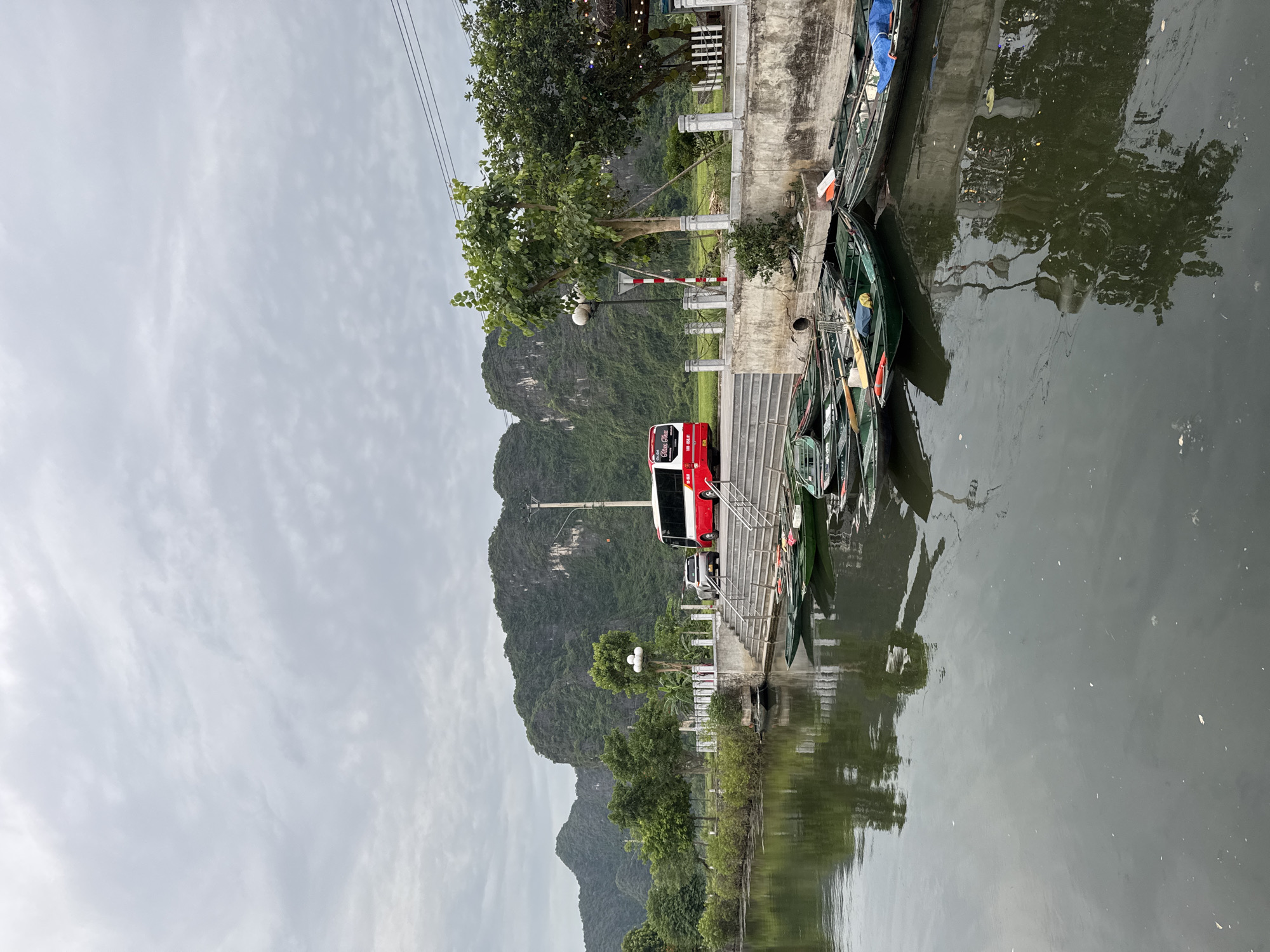 A grey, cloudy sky hangs over a waterfront scene in Nam Hoa Lư Ward, Vietnam. In the foreground, a calm, dark body of water reflects the overcast sky and the trees lining the opposite bank. Several small, green, rowboat-like vessels are moored along the bank, some with blue tarps covering parts of them. A concrete walkway with white railing runs along the bank, elevated slightly above the water.
On the walkway, a red and white bus is parked, facing left, with a silver car behind it. The bus has "Hao Vien" written on its side. A series of concrete steps leads down from the walkway towards the water.
In the background, a lush, green mountain range with jagged peaks rises dramatically. Trees and smaller vegetation are scattered across the landscape. A tall, thin light pole stands on the walkway, and power lines are visible in the upper right corner of the frame. The overall impression is one of natural beauty with signs of human activity and transportation.