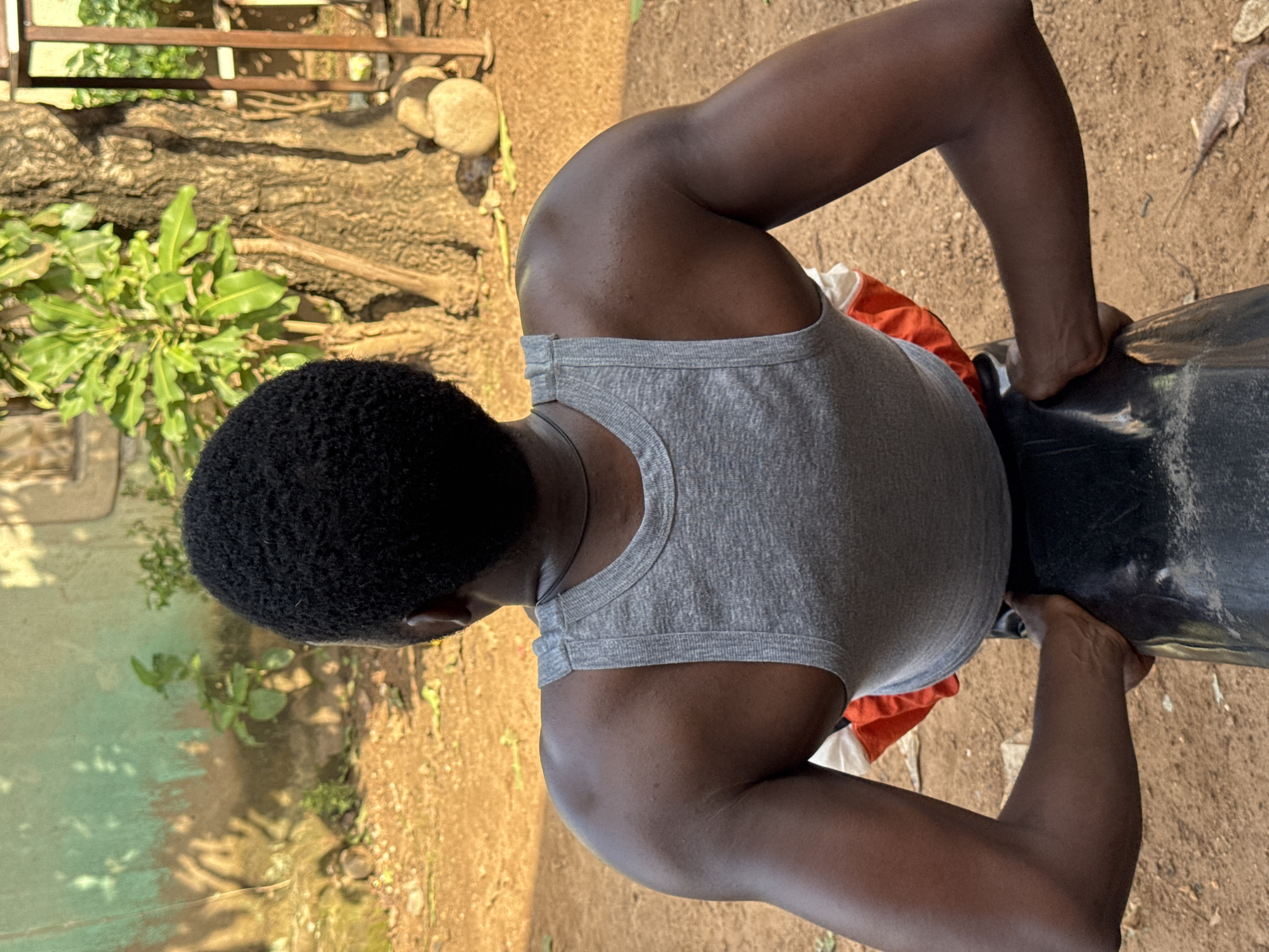 The image shows a person from behind, wearing a grey tank top and orange and white shorts. The person has dark skin and short, curly black hair. They are seated on a black object, possibly a chair or bench, with their arms resting on it. The background appears to be an outdoor setting with dirt ground, a tree, and a building with a green wall. Sunlight casts shadows on the ground and wall. Based on the visual cues, this scene is likely located in Akwanga, Nigeria.