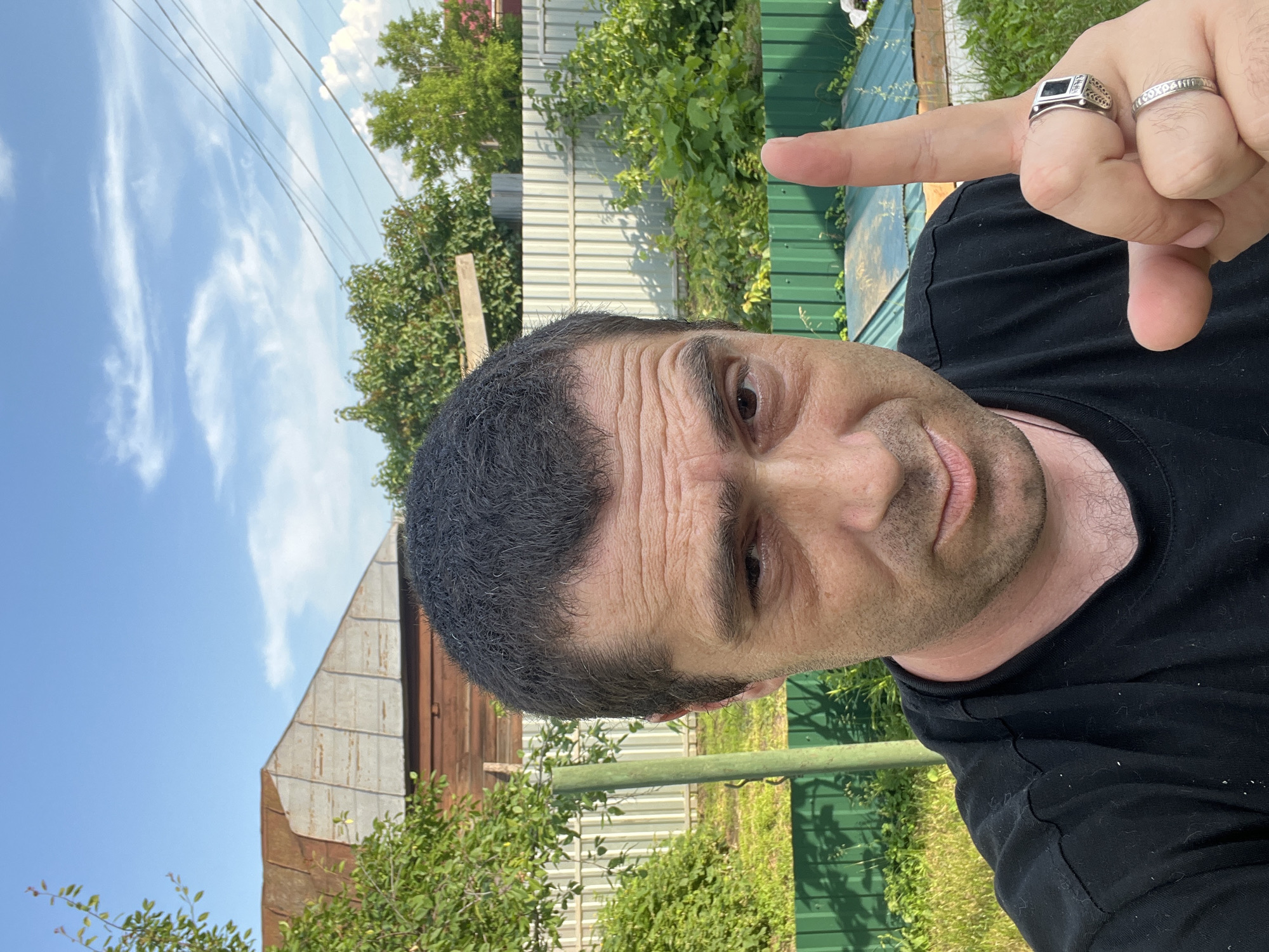 A close-up, medium shot shows a Caucasian man in his late 30s or early 40s with dark, short hair and brown eyes, looking directly at the camera with a slight smile. He is wearing a black t-shirt and a silver ring with a black stone on his left ring finger. His right index finger is raised, pointing upwards. The man is outdoors, with a blurred background of a wooden house and a green fence, suggesting a rural or suburban setting. The sky is blue with scattered clouds, and there are green trees and bushes visible. The lighting suggests it is daytime, possibly late afternoon due to the angle of the shadows.