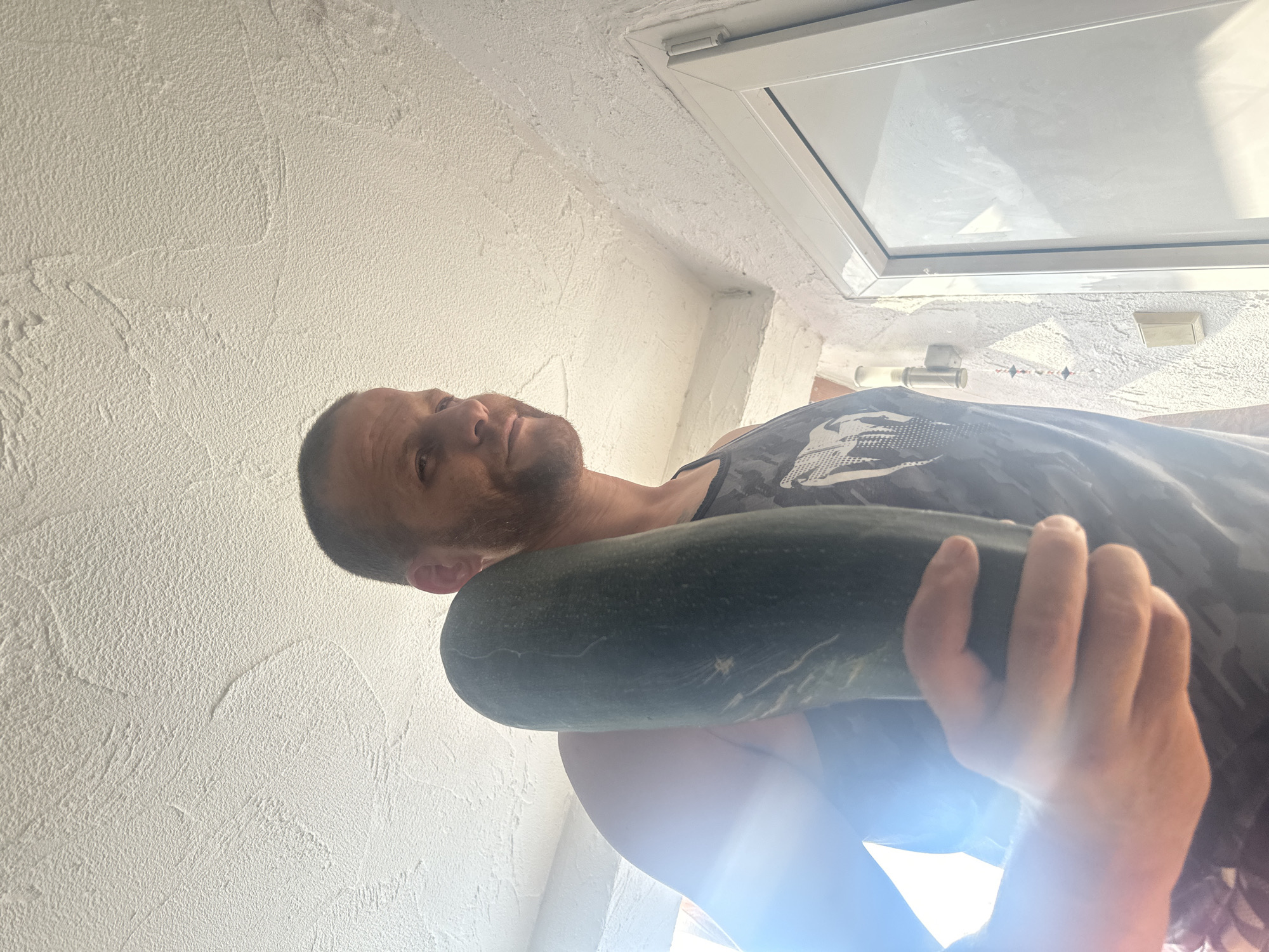 A man, approximately 40-50 years old, with short brown hair and a beard, stands indoors, holding a large zucchini in his right hand. He is wearing a dark-colored tank top with a graphic print. The background features a textured white ceiling and wall, and a white door frame. Sunlight streams in from the left, creating lens flare and illuminating the scene. The image appears to be taken from a low angle, looking up at the man. The environment suggests a domestic setting, possibly a home or a covered outdoor area, in Heinebach, Germany.
