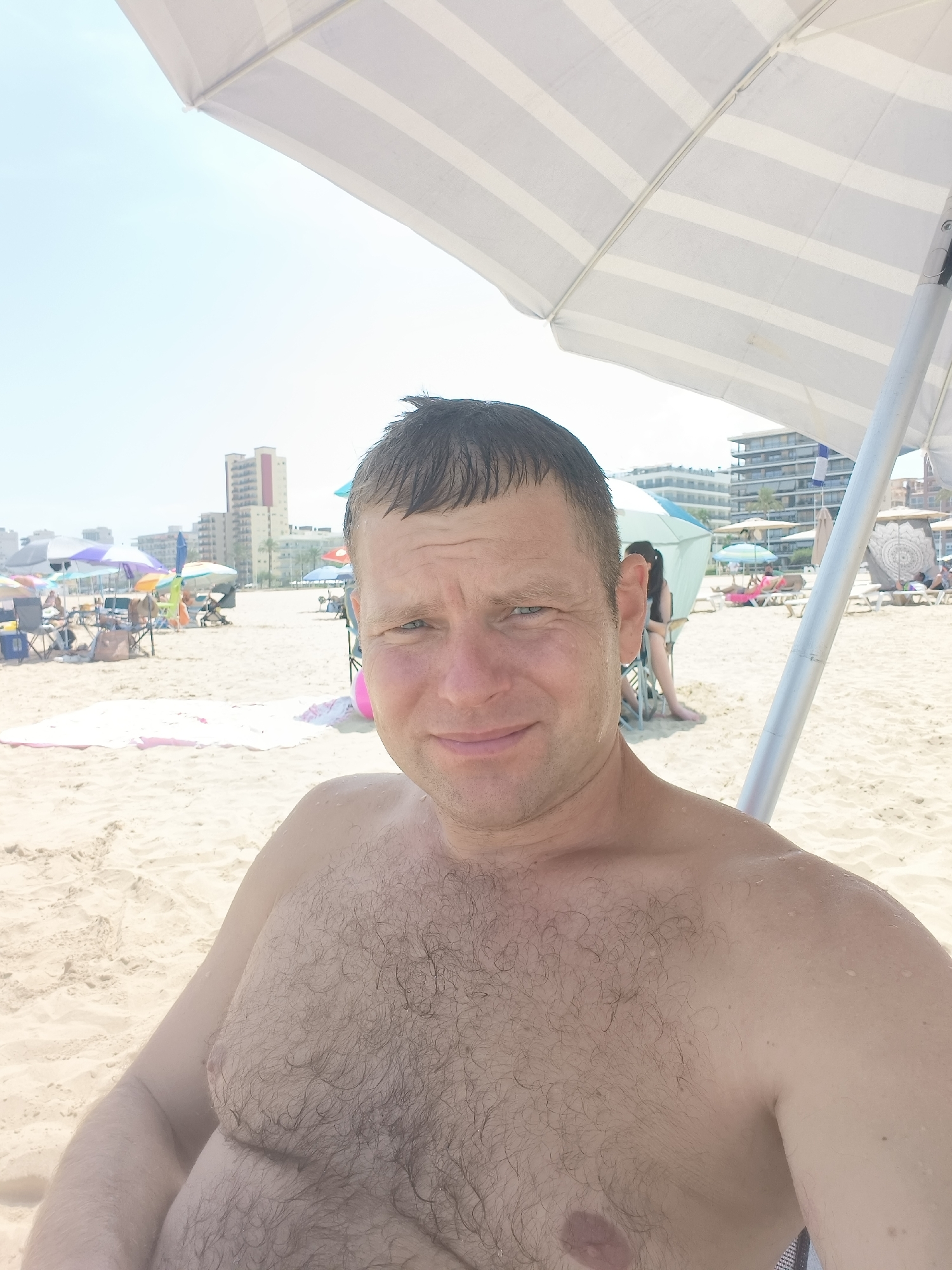 A man with short, dark hair and a hairy chest sits on a sandy beach in Gandia, Spain, under a striped umbrella. He is looking at the camera with a slight smile. In the background, there are other beachgoers, umbrellas, chairs, and buildings. The sky is clear and blue.