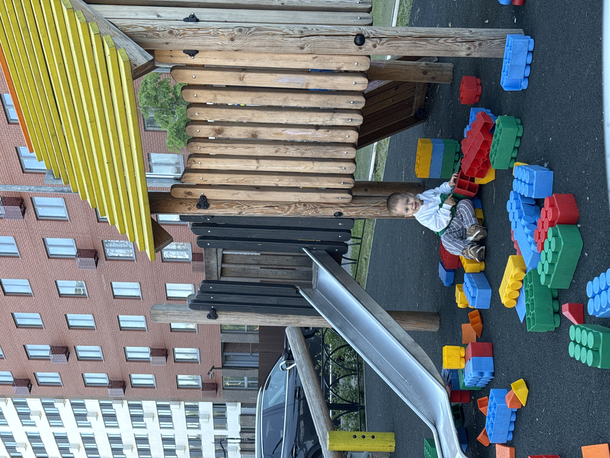 This image depicts an outdoor playground scene in Tomilino, Russia, featuring a young child playing amidst large building blocks.
The primary subject is a smiling toddler or preschooler, with light hair, seated on a dark, rubberized playground surface. The child is wearing a white long-sleeved top with green details, striped pants, and sneakers, and looks directly at the camera with a happy expression.
The child is surrounded by numerous large, colorful interlocking building blocks in blue, red, green, and yellow, which are scattered across the ground, some forming small structures.
In the mid-ground, a robust wooden playground structure is prominent. It features a distinctive yellow slatted roof or awning, wooden walls resembling a playhouse or fort, and a silver-colored slide extending from one side. A black bench or seating area is integrated into the wooden framework.
The background reveals multi-story residential buildings constructed from red brick, featuring numerous windows. Some windows have external units, possibly air conditioners or planter boxes. A green tree provides natural foliage behind the wooden structure. A dark-colored car is partially visible parked near the playground.
The scene takes place during the daytime under soft, diffuse lighting, suggesting an overcast sky or a shaded area. No visible text is discernible in the image.