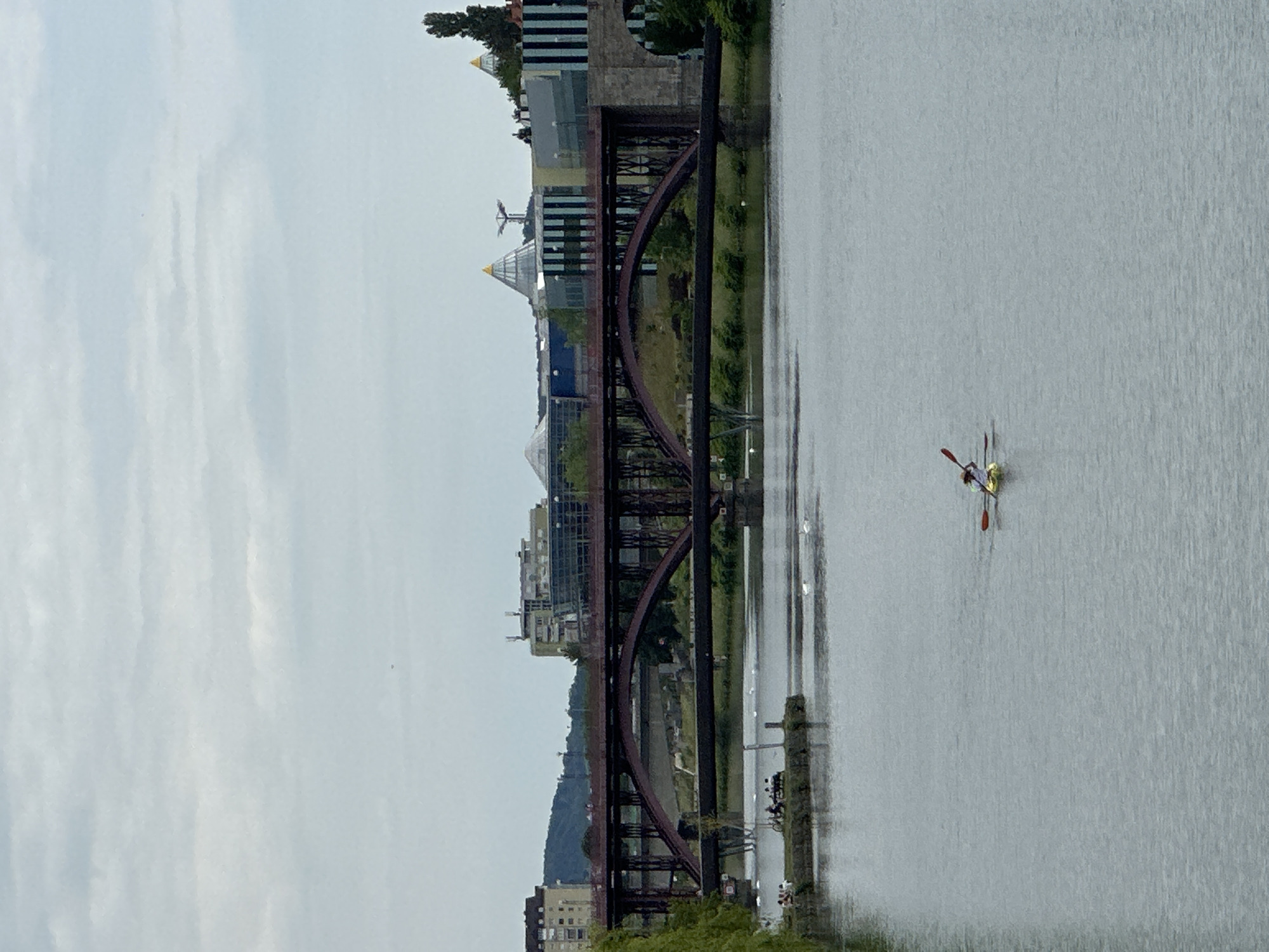 The image shows the Drava River in Maribor, Slovenia, with the Old Bridge (Stari most) spanning the river. The bridge is a metal structure with arched supports. Modern buildings with pyramidal roofs are visible behind the bridge. A single scull rowing boat with a person is on the river's surface. The sky is overcast.