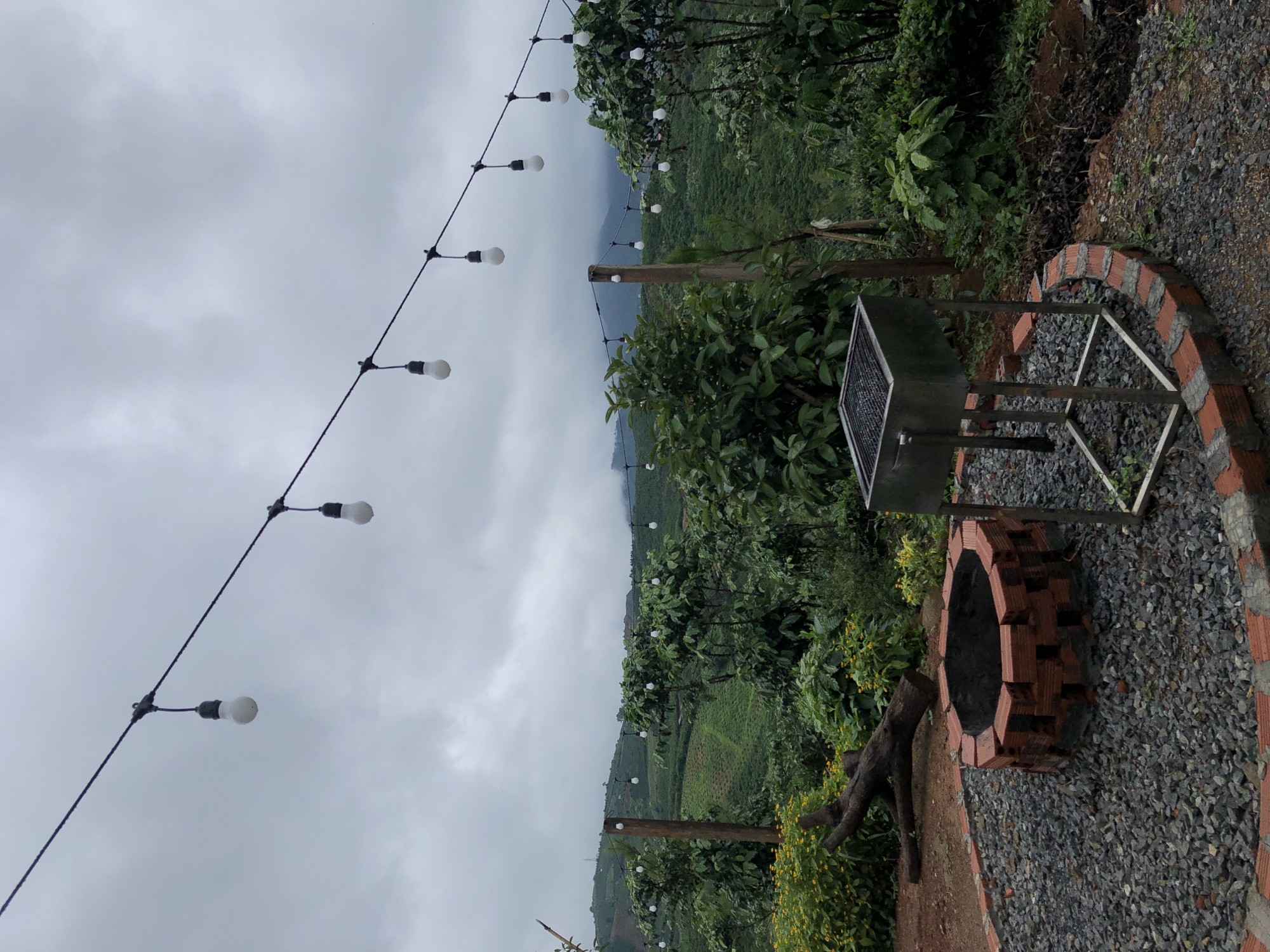 The image depicts an outdoor barbecue area at a rural location, likely in Thôn 4, Vietnam. The scene is characterized by a cloudy, overcast sky that casts a diffused light over the landscape. In the foreground, a circular brick fire pit, filled with grey gravel, is situated next to a metal barbecue grill. A weathered wooden log lies on the gravel nearby. Surrounding the barbecue area is lush green vegetation, including trees and bushes, with a backdrop of rolling hills covered in agricultural terraces, suggesting a coffee or tea plantation. A string of decorative light bulbs hangs across the scene, suspended from a wire, adding to the ambiance of the outdoor space. The overall impression is one of a serene, natural setting, prepared for outdoor gatherings.