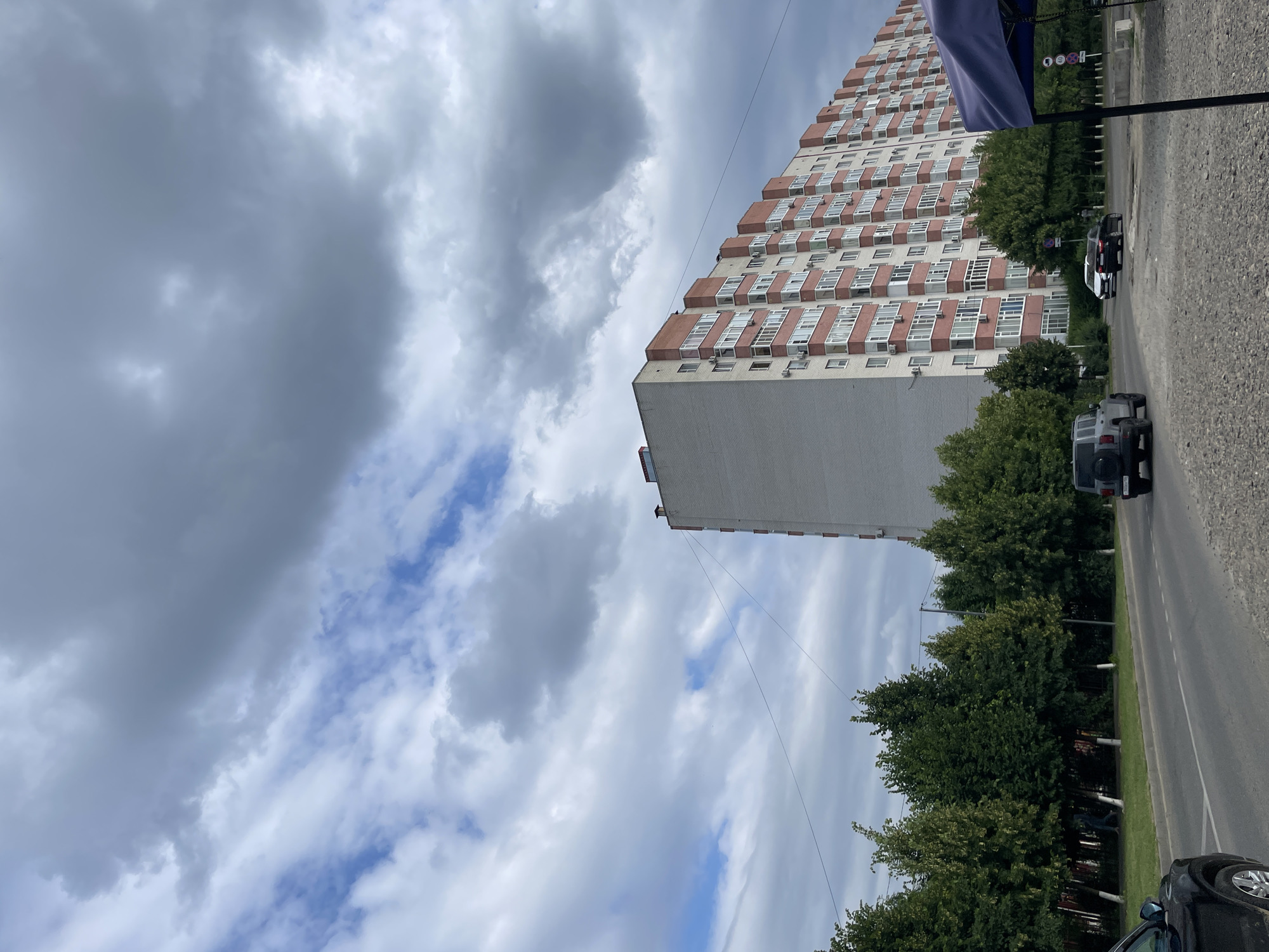 In Stavropol, Russia, a multi-story apartment building with a white brick facade and red balconies dominates the skyline under a cloudy sky. A line of trees with white-painted trunks runs along the street in front of the building. Several cars are visible on the road, including a dark SUV, a black sedan, and a white sedan. A dark-colored car is parked on the left side of the frame, and a blue canopy is partially visible in the lower right corner. The ground is covered with gravel.