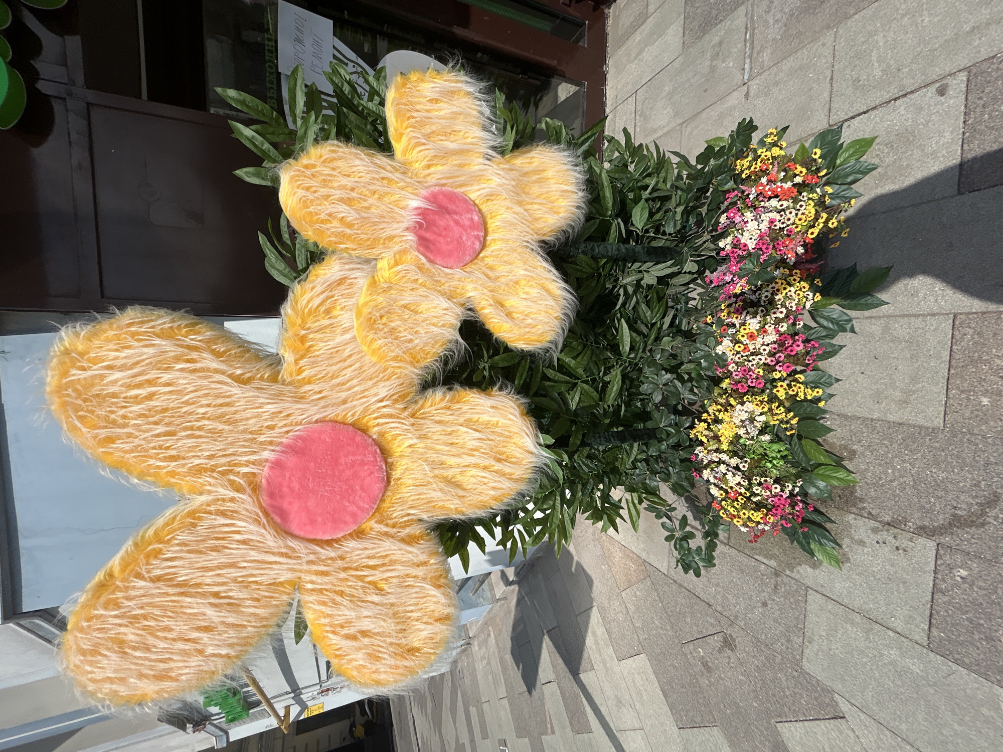 Two large, oversized yellow fabric flowers with pink centers are positioned next to a bush with small, multi-colored flowers, outside a building. The scene appears to be on a city street, with grey stone paving visible. The location is Unknown, Unknown.