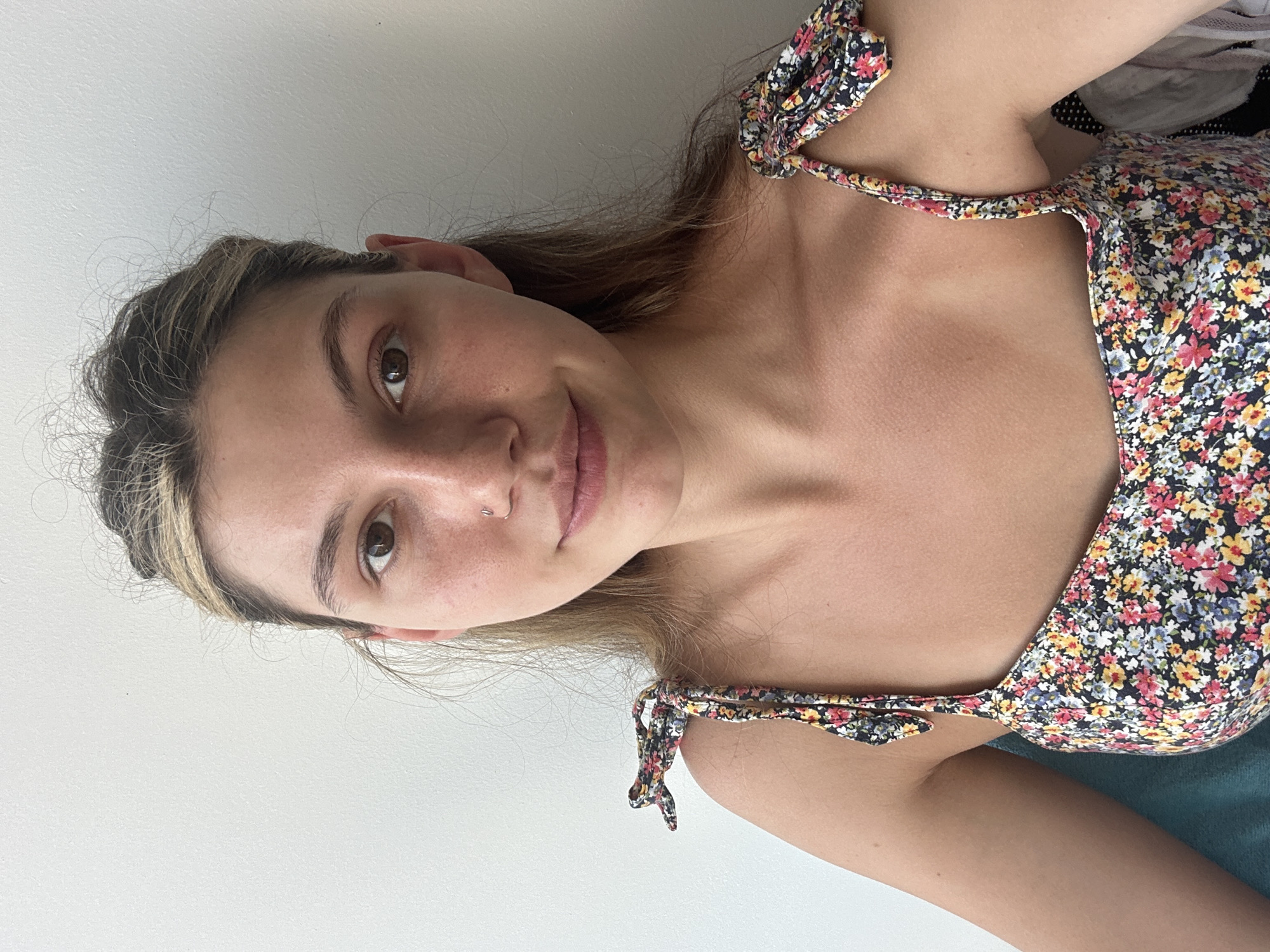 A young woman is shown in a close-up shot, likely a selfie, against a plain white wall. She has brown hair with blonde highlights, brown eyes, and a nose piercing. She is wearing a floral print top with tie straps. The background is out of focus, but there's a hint of fabric or clothing, possibly a pile of clothes. No evidence suggests the location is Baan Chaweng, Thailand.