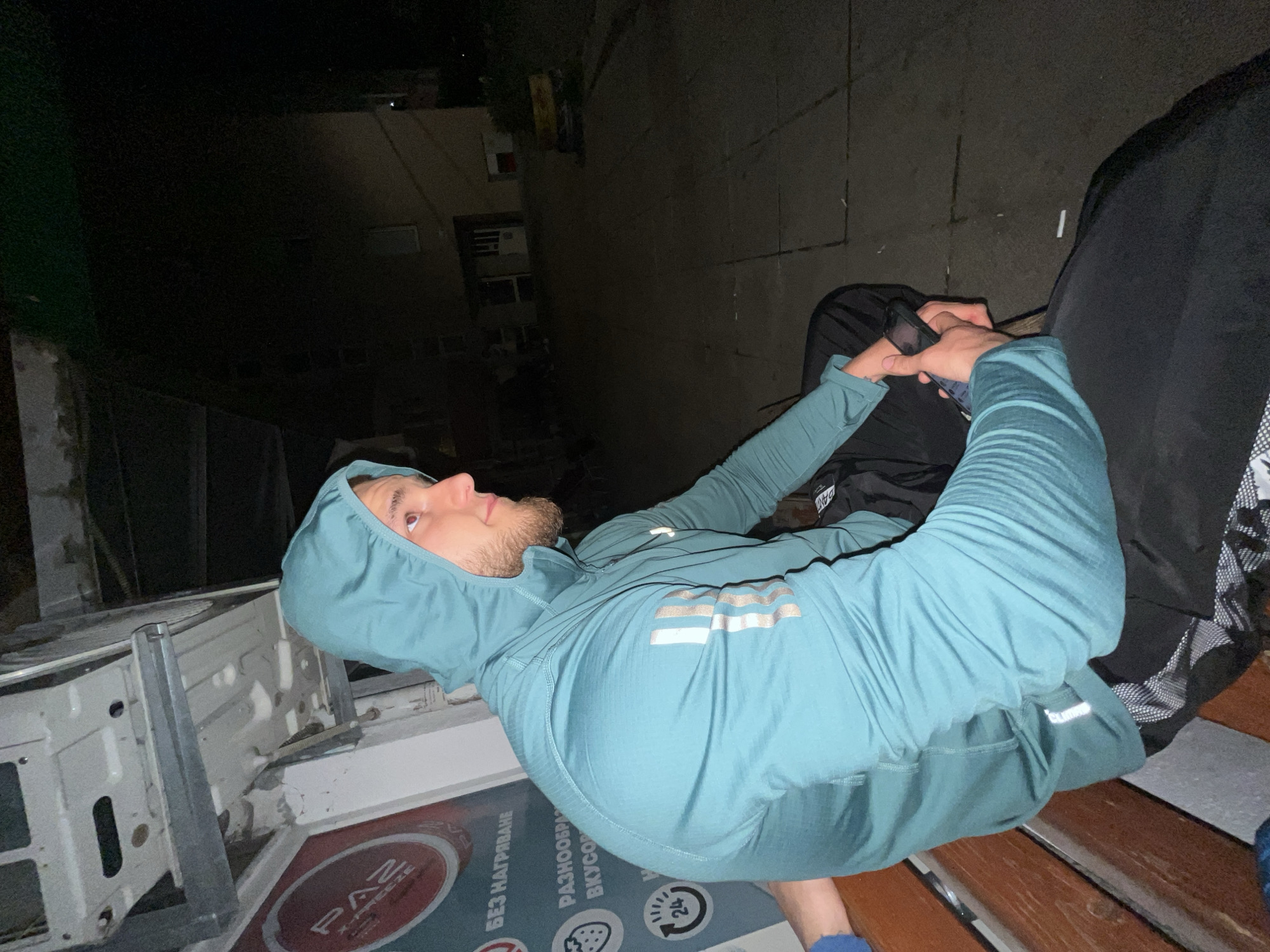 A man wearing a turquoise hooded tracksuit sits on a wooden bench at night, holding a smartphone in his hands. He has a beard and is looking upwards. The bench is located near a building with a commercial sign in Bulgarian, indicating "PAZ". Behind the bench is a dark alleyway, with buildings visible in the distance. The location appears to be in an urban setting, potentially within the city of Burgas, Bulgaria.