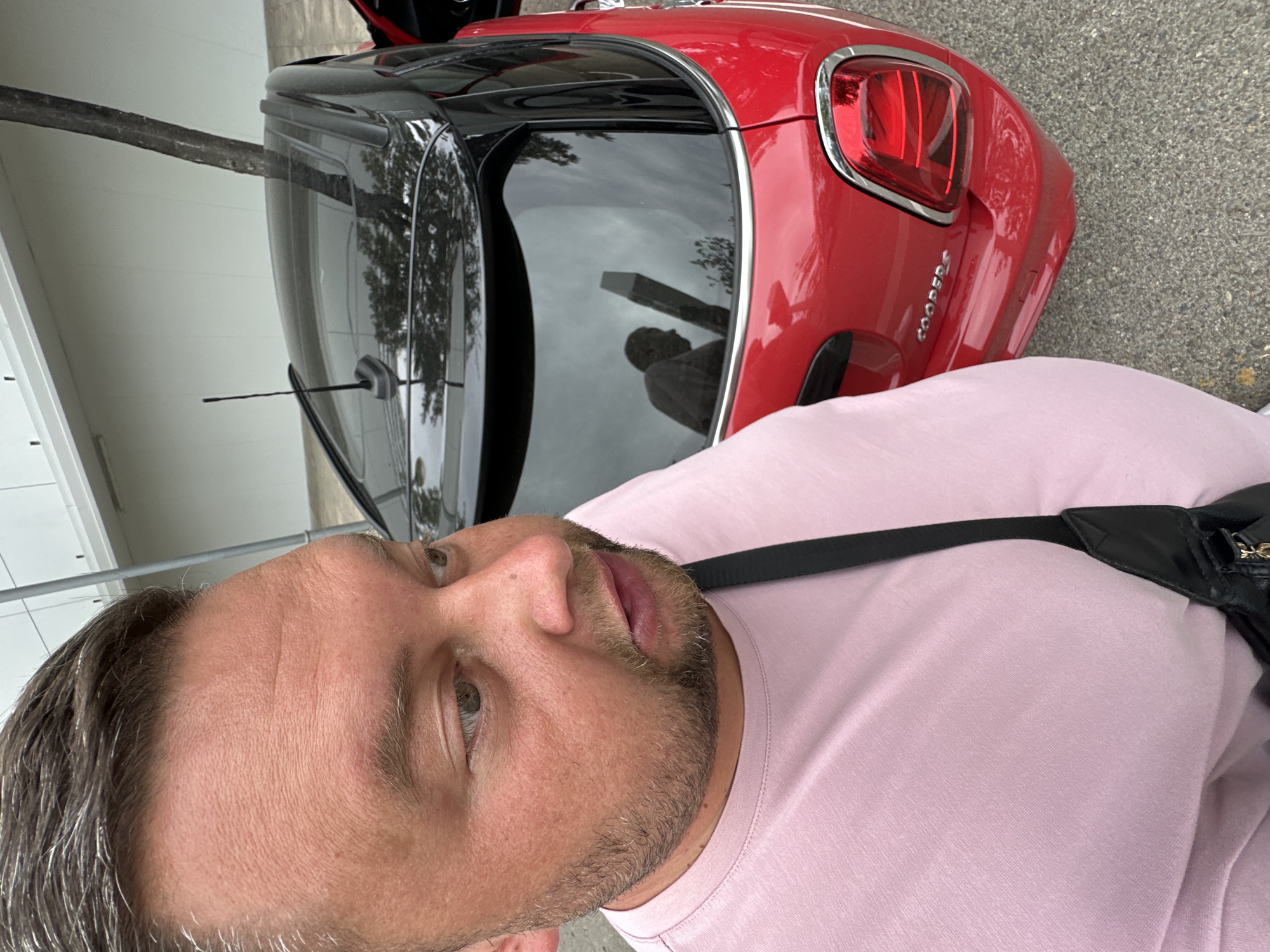 A man wearing a pink t-shirt and a black crossbody bag is standing next to a red Mini Cooper. The car has a black roof and a black antenna. The man's face is partially visible in the foreground, with his left eye looking to the right. The car's rear window reflects a person and a tree. The scene appears to be outdoors, possibly in a parking lot or street, with a light-colored building and a tree visible in the background. The ground is asphalt or pavement.