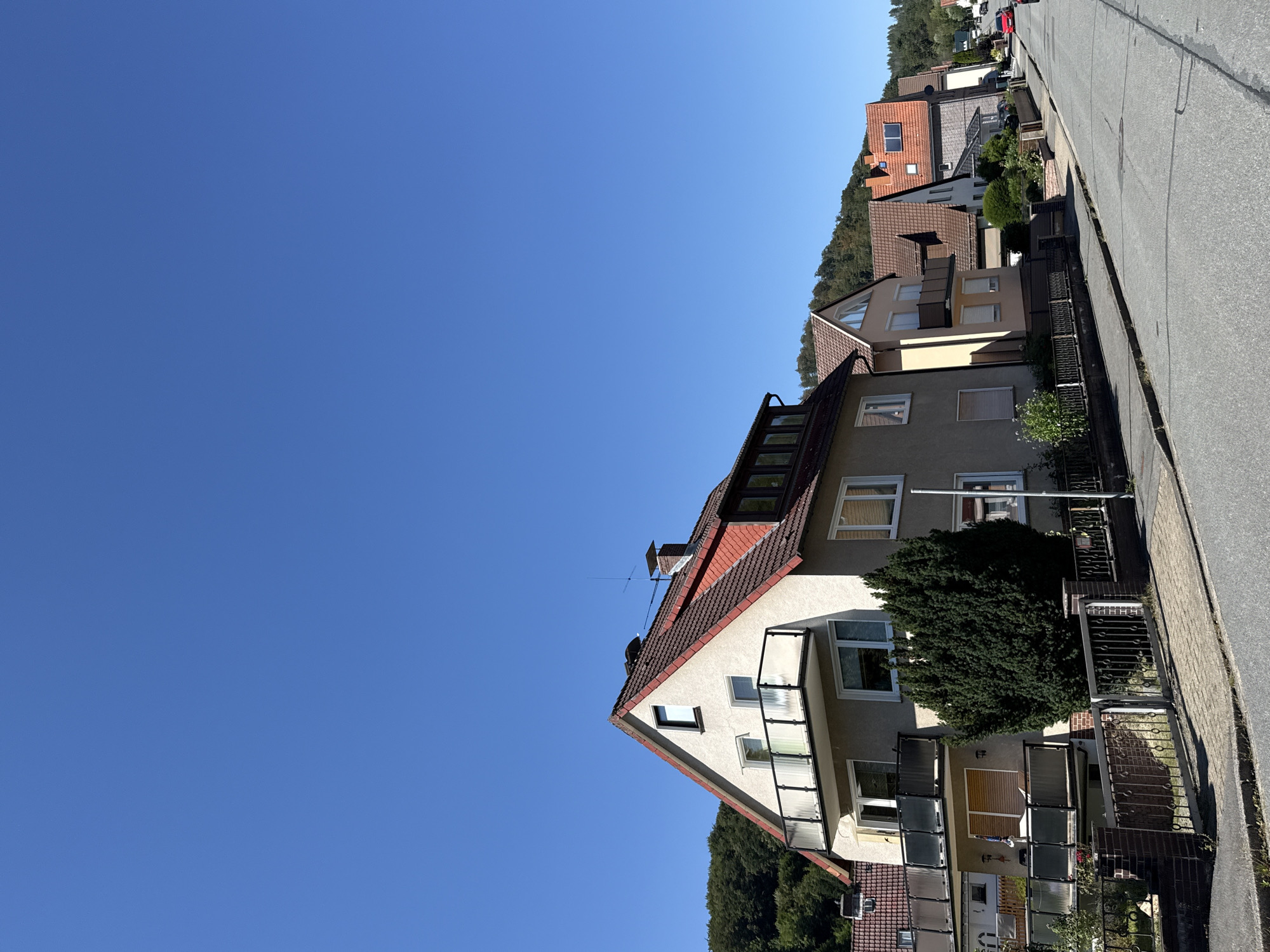 The image shows a street in the city of Bad Lauterberg im Harz, Germany. The street is lined with houses, and there is a clear blue sky overhead. The houses appear to be residential, with multiple stories and balconies. Trees and foliage are visible on the hills in the background. The street is paved and there are sidewalks on either side. A red car is visible in the distance on the right side of the image. The image appears to have been taken during the daytime on a sunny day. There are no people visible in the image.