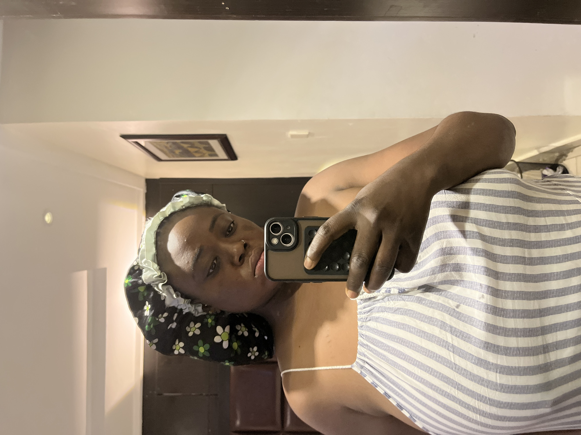 A woman is taking a selfie in a room. She is wearing a bonnet and a striped garment. She is holding an iPhone. A picture frame is visible on the wall. There is no information to conclude that this picture was taken in Abuja, Nigeria.