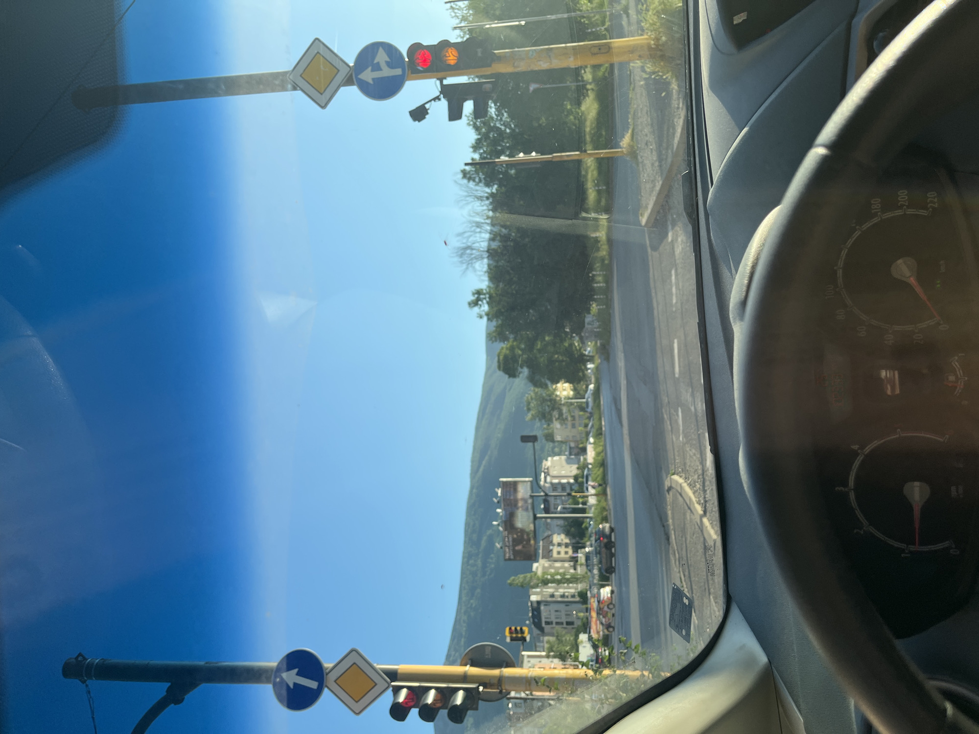 The image shows a view from inside a vehicle, likely a car, looking out onto a street in the city of Unknown, Unknown. The sky is clear blue, and the sun is shining. In the foreground, the interior of the car is visible, including the dashboard, steering wheel, and a portion of the windshield. Traffic lights are visible through the windshield, both near and far. The closer traffic lights are mounted on yellow poles with road signs indicating a primary route ahead and that the left turn has the right of way. Further down the road, buildings and trees line the street, with a mountain visible in the background. The scene suggests a sunny day in an urban environment near a natural landscape.