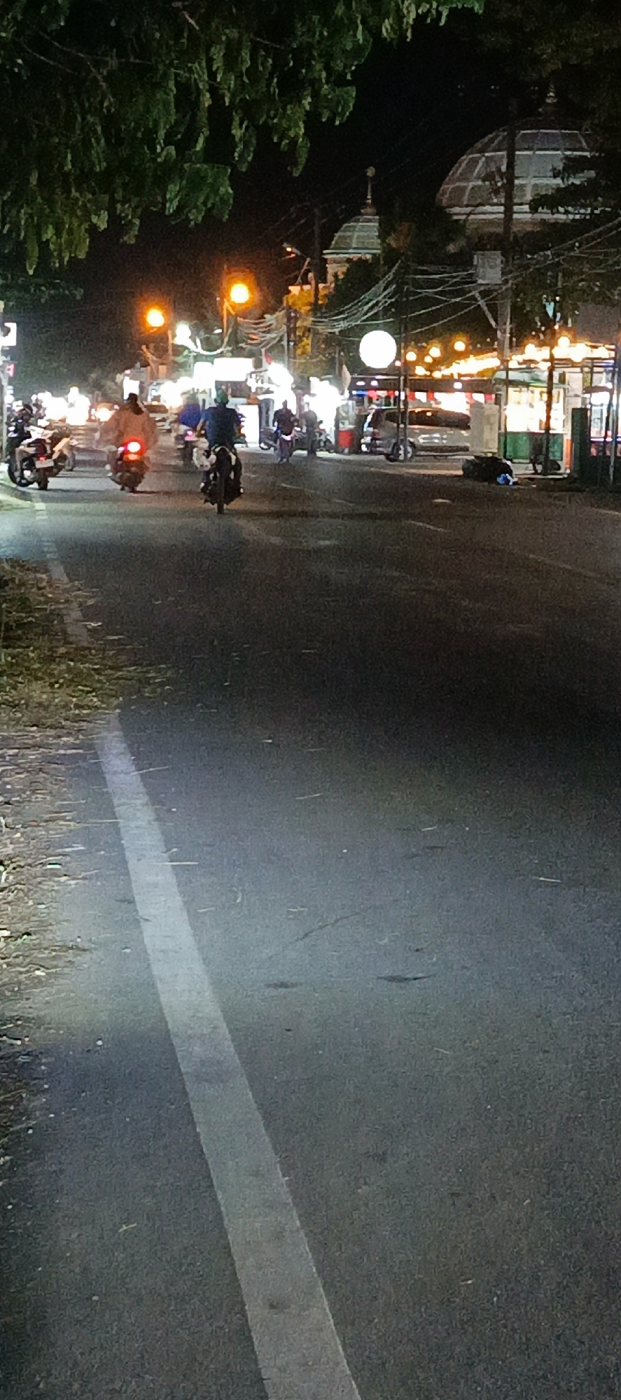 This is a nighttime, vertical, full shot of a road in Banda Aceh, Indonesia. The road is paved with asphalt and has a white line painted on the left side, indicating a lane divider. The road is wet, reflecting the surrounding lights.
On the right side of the road, there are several stalls or shops illuminated by bright lights. In front of these stalls, there are some people and a car. A dark, lumpy object, possibly trash or luggage, is on the ground near the stalls.
Several motorcycles are visible on the road. One motorcycle in the foreground has a rider wearing a dark shirt and helmet, moving away from the camera. Further down the road, other motorcycles and riders are seen, some with their headlights on, creating streaks of light.
Above the road, there are trees with dark green leaves. In the background, the distinctive domed architecture of a building, likely a mosque, is visible, illuminated by lights and decorations strung across the street. Streetlights are also present, casting a warm glow. The overall scene suggests a busy, commercial area at night.
