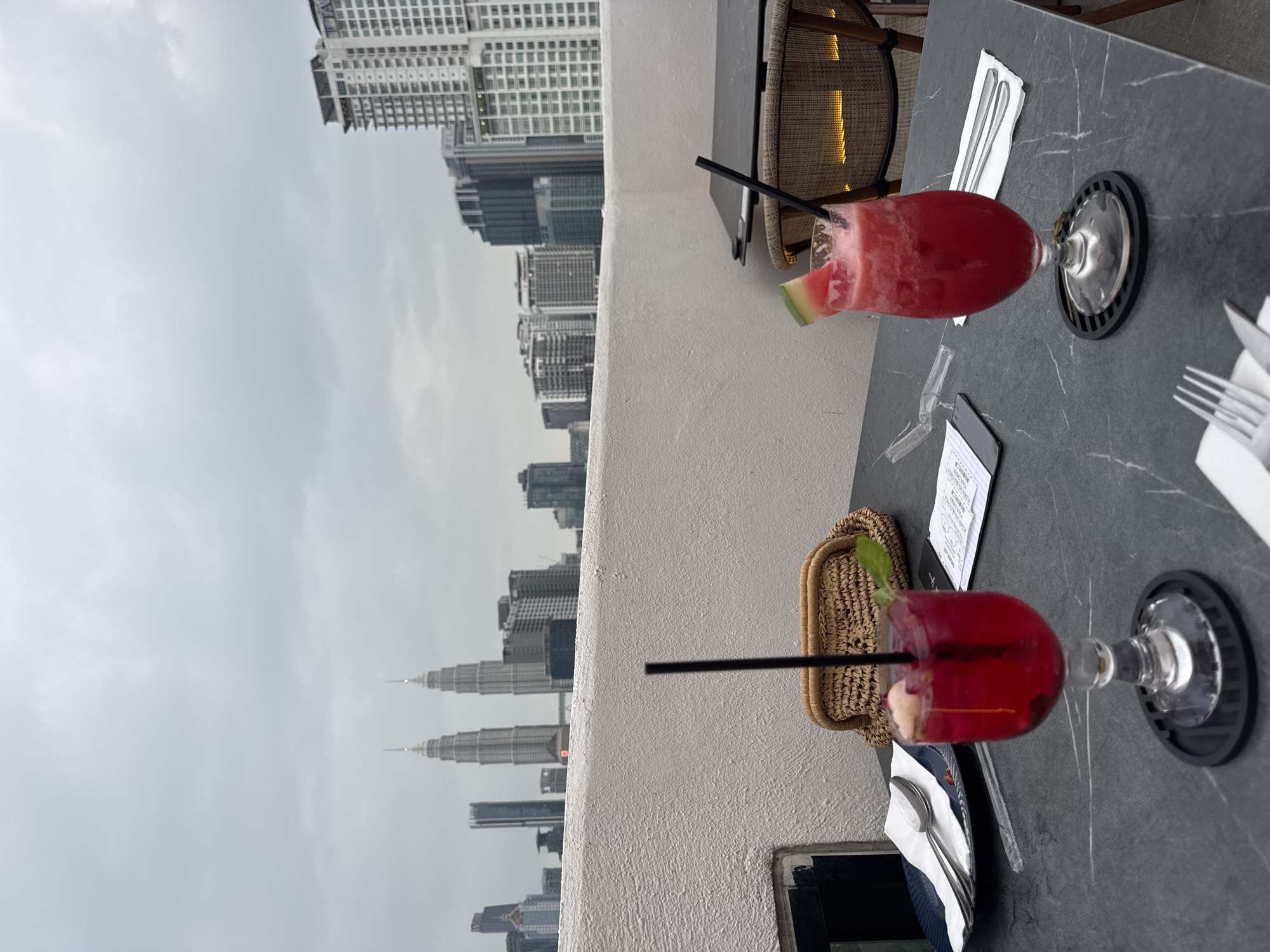 This image shows a rooftop scene in Kuala Lumpur, Malaysia, featuring a table set for two with drinks. In the background, the Petronas Twin Towers can be seen among other buildings in the city skyline. Two stemmed glasses containing red beverages with straws are placed on the table. The drink on the left has a slice of watermelon as garnish. Next to each drink, there's a set of cutlery wrapped in a white napkin. A small woven basket and a black menu card are placed on the table. The weather appears overcast with a grey sky. The table is made of dark grey material with a textured pattern. A rattan chair is partially visible at the edge of the table.