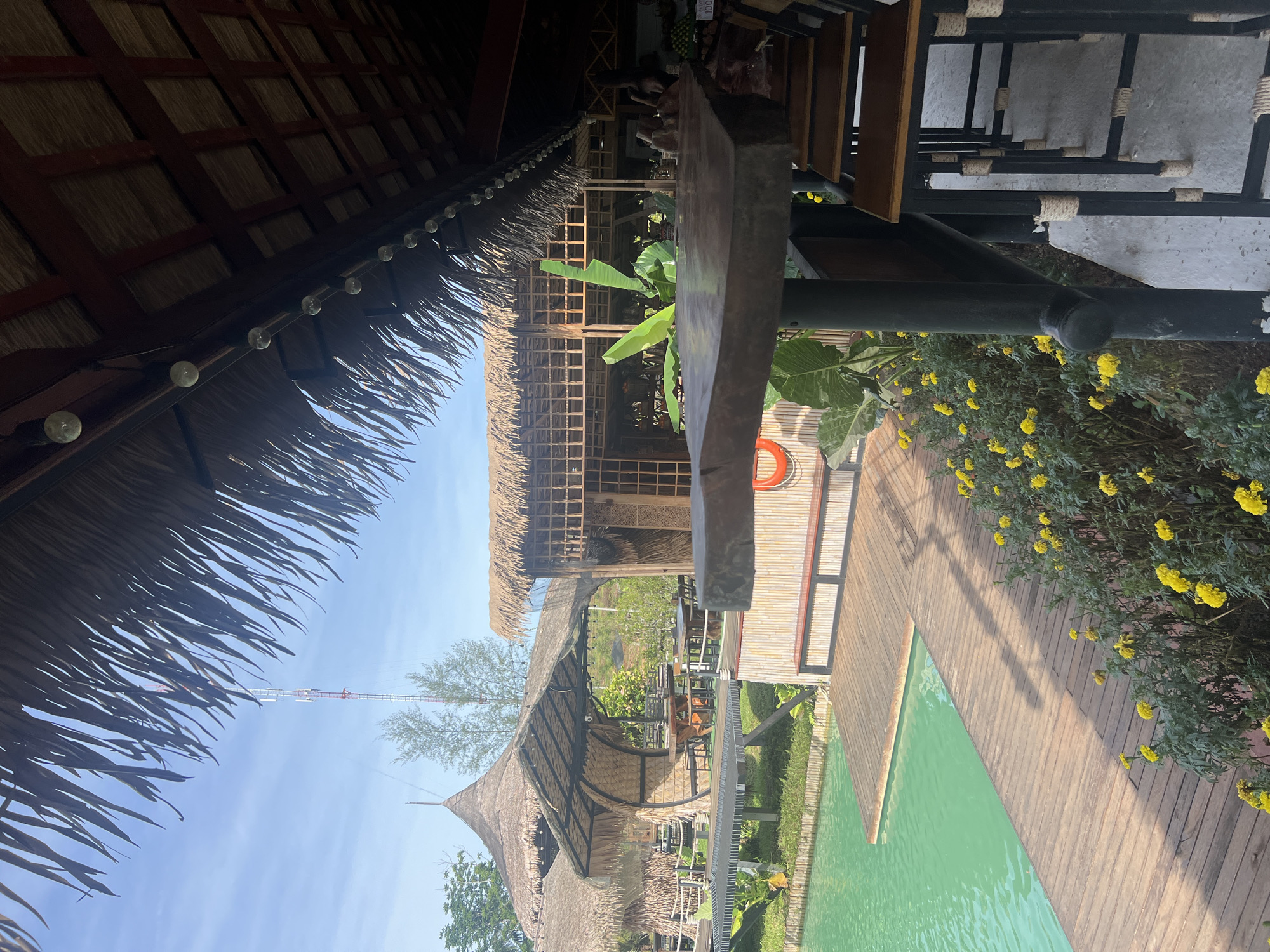 This is a medium-shot, eye-level, outdoor photograph of a restaurant with a tropical ambiance, located in Tambon Pa Kae Bo Hin, Thailand. The scene features a thatched-roof structure with an open-air seating area overlooking a vibrant turquoise body of water. Wooden decking extends from the restaurant towards the water, with a wooden railing in the foreground. Several bar stools with wooden seats and black metal frames are arranged at a high wooden table. Yellow flowers are blooming in the foreground, adding a splash of color. In the background, additional thatched-roof structures are visible, along with greenery and a distant telecommunications tower. The sky is partly cloudy and blue. The overall impression is one of a relaxed, scenic dining environment.