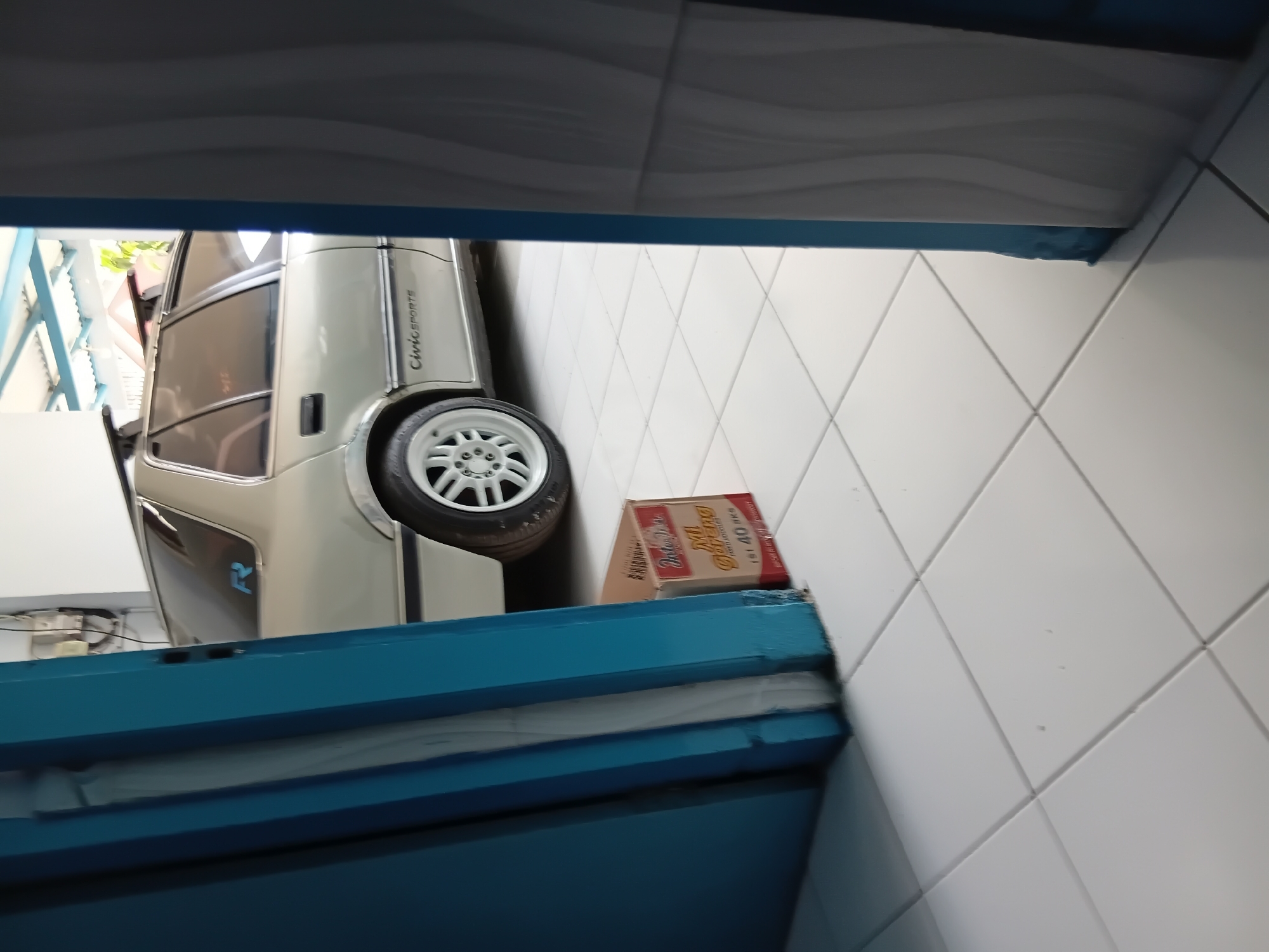 An outdoor shot shows a silver sedan parked on a tiled floor, partially obscured by a blue doorway. The car has a roof rack and the words "Civic SPORTS" are visible on its side. A cardboard box of "Mi Goreng" instant noodles sits on the floor near the car. The background reveals a corrugated roof structure and some greenery. The image was taken in Tanggul Wetan, Indonesia.