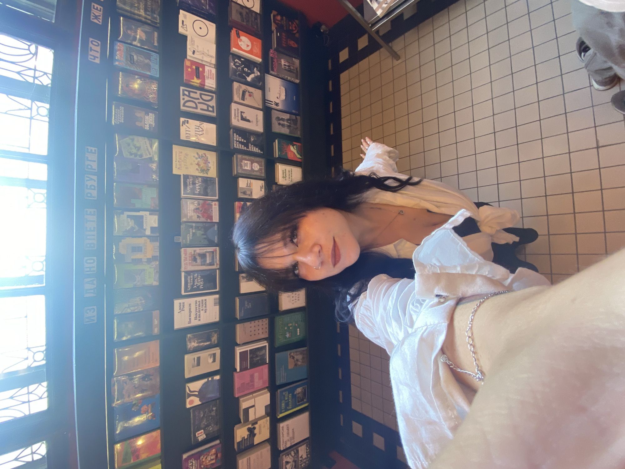 A young woman with dark hair and bangs, wearing a white blouse and a necklace, poses for a selfie in a bookstore. She is positioned in the foreground, with rows of books lining the shelves behind her. The store appears to have a tiled floor and a ceiling with decorative windows. The woman's left arm is extended towards the camera, and her right arm is resting on a shelf.