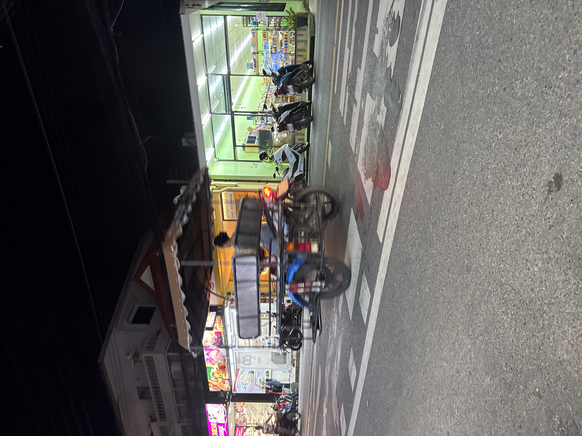 In the city of Karon, Thailand, at night, a motorized tricycle with a passenger is seen driving on a road. The tricycle is a three-wheeled vehicle with a covered passenger area. The road has a pedestrian crossing with white stripes. On the right side of the road, a brightly lit store with green windows is visible, stocked with shelves of goods. Several motorcycles are parked outside the store. On the left side of the road, a building with balconies and awnings is seen, with neon signs displaying images. The overall scene depicts a bustling street in Karon at night, with commercial activity and local transportation.