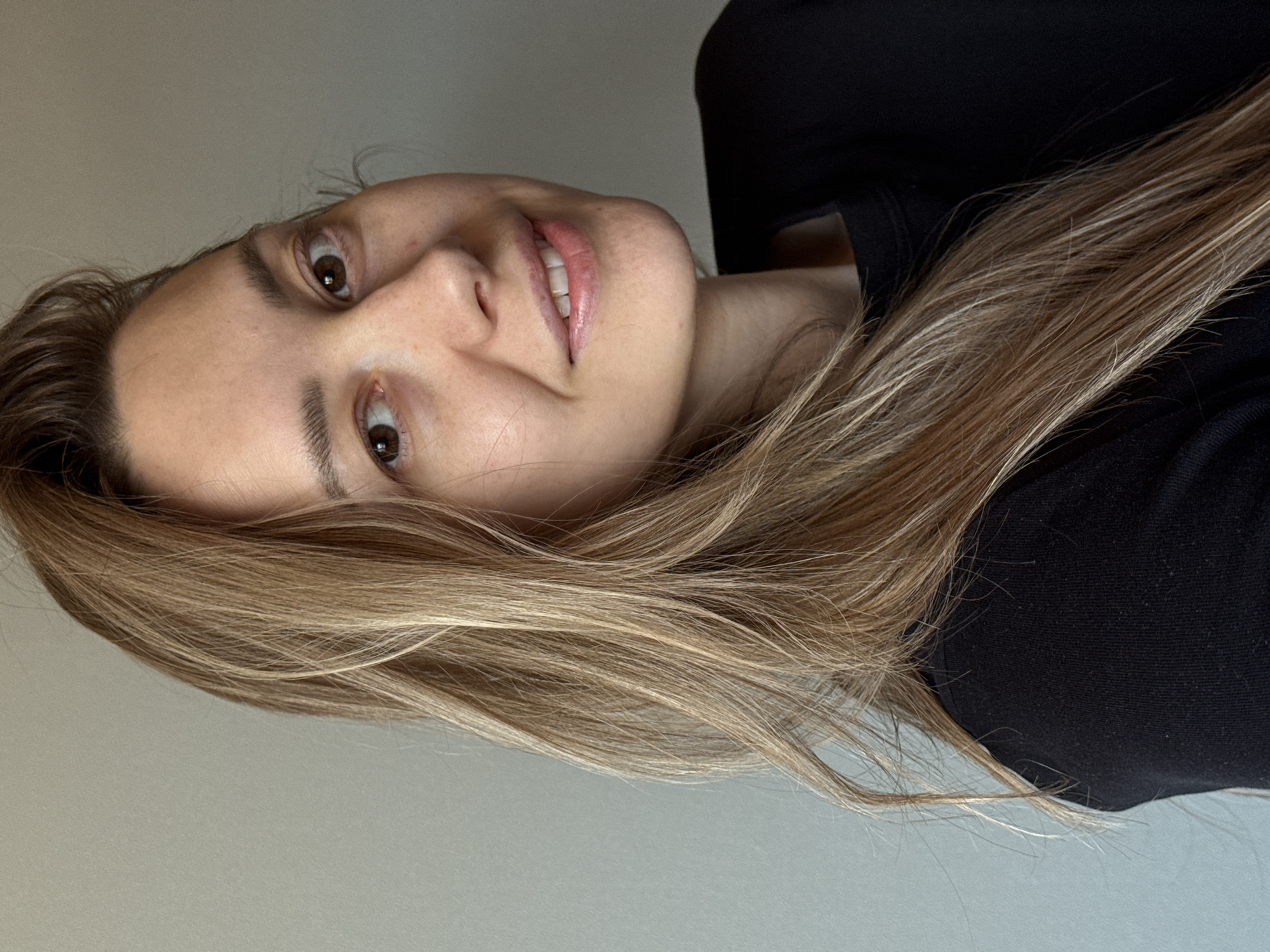 A head-and-shoulders portrait captures a young adult woman looking directly at the camera with a gentle, slight smile. She has warm brown eyes and fair skin. Her long, straight or slightly wavy hair, in shades of light brown and blonde, frames her face and falls over her shoulders, covering parts of the frame. She is dressed in a dark, likely black, long-sleeved top with a simple neckline. The background is a plain, light, neutral-colored wall, indicating an indoor setting. The lighting is soft and even, suggesting either natural light indoors or well-distributed artificial light, casting minimal shadows. Her expression is calm and approachable. There are no visible objects other than the woman and her attire, and no text is present in the image. The image itself does not contain specific visual cues to indicate the given location of Chaikovsky, Russia.