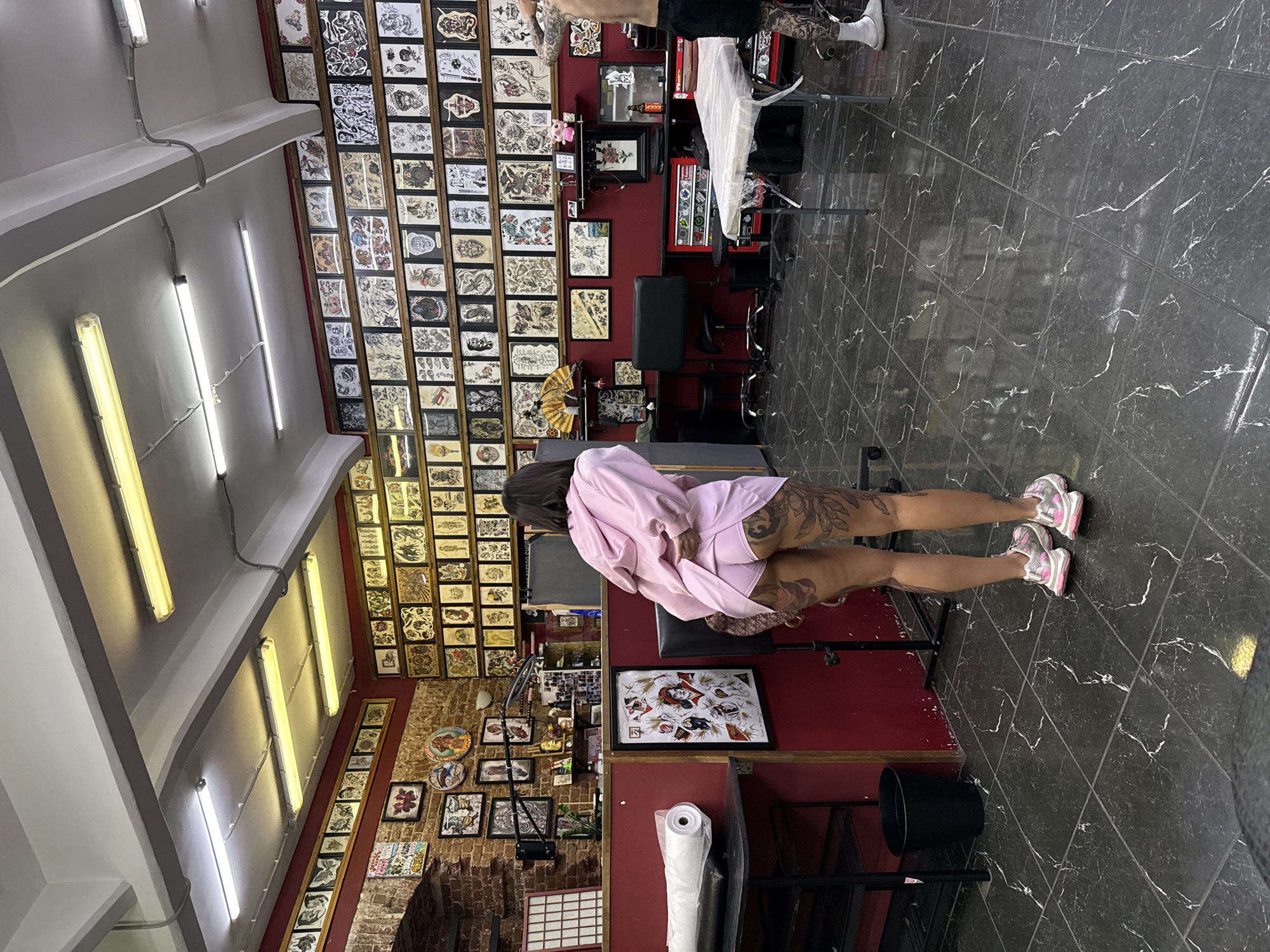 A person sits in a tattoo parlor. The subject is female with short black hair. She wears a pink long-sleeved garment, and shorts of the same color. The subject wears white and pink sneakers. Tattoos are visible on her upper legs. She sits on a black leather tattoo chair. The floor is dark gray marble. The walls are lined with tattoo flash sheets. There are fluorescent lights on the ceiling. There is a black garbage can near the subject's right foot. Based on the appearance of the tattoo flash sheets and the brickwork, this is likely a studio located in Moscow, Russia. Another person is partially visible standing near the top right of the photo. They are getting a tattoo on their thigh, and it is in progress.