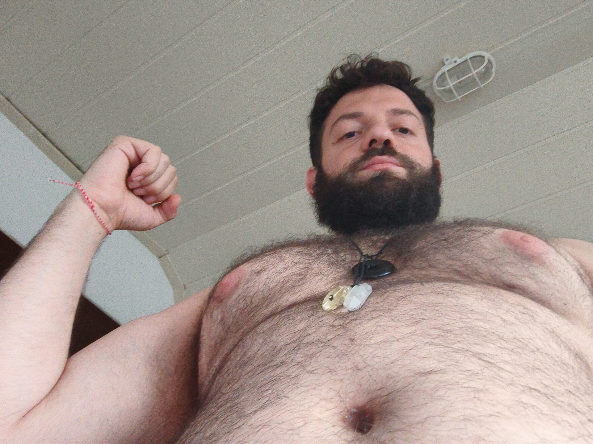 A Caucasian man with a dark beard and hairy chest looks down at the camera. He is wearing a necklace with several pendants. His left arm is flexed, and a red and white string bracelet is visible on his wrist. The background is a white ceiling with a caged light fixture. The image does not contain any information that would identify its location as Diepholz, Germany, or any other specific location.