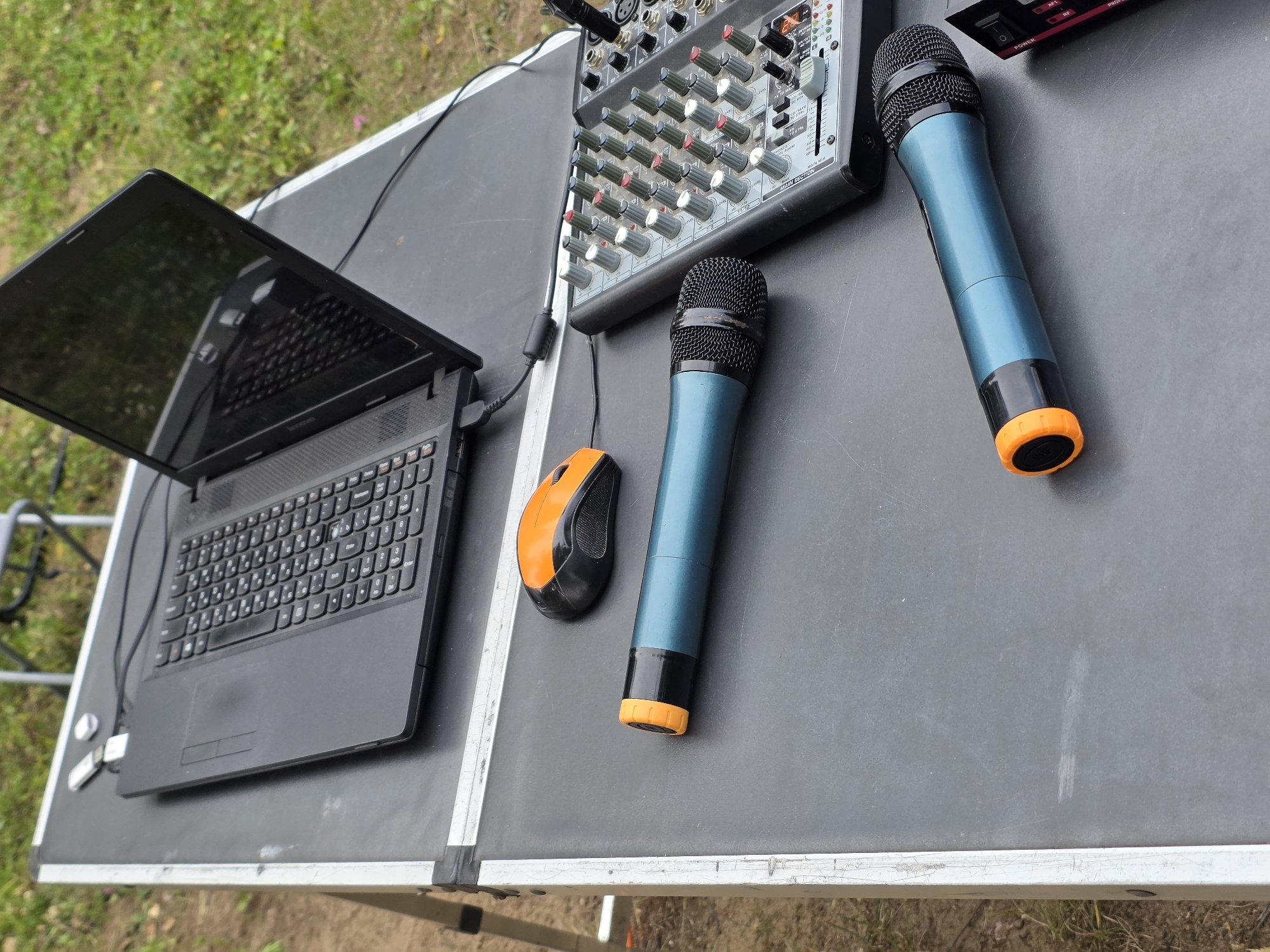 The image depicts an outdoor setup for audio equipment, potentially for an event or performance. A black laptop is open on a large, dark-gray case. Next to the laptop, there is an orange and black computer mouse. Two teal-colored microphones with orange accents are lying on the case, one in the foreground and another further back. To the right of the microphones is a sound mixer with numerous knobs and sliders. The case is resting on what appears to be a metal stand, and the background shows green grass and foliage, suggesting a park or outdoor venue. The specific location in Chaikovsky, Russia, is not evident from the visual information alone. There are no people visible in the image.