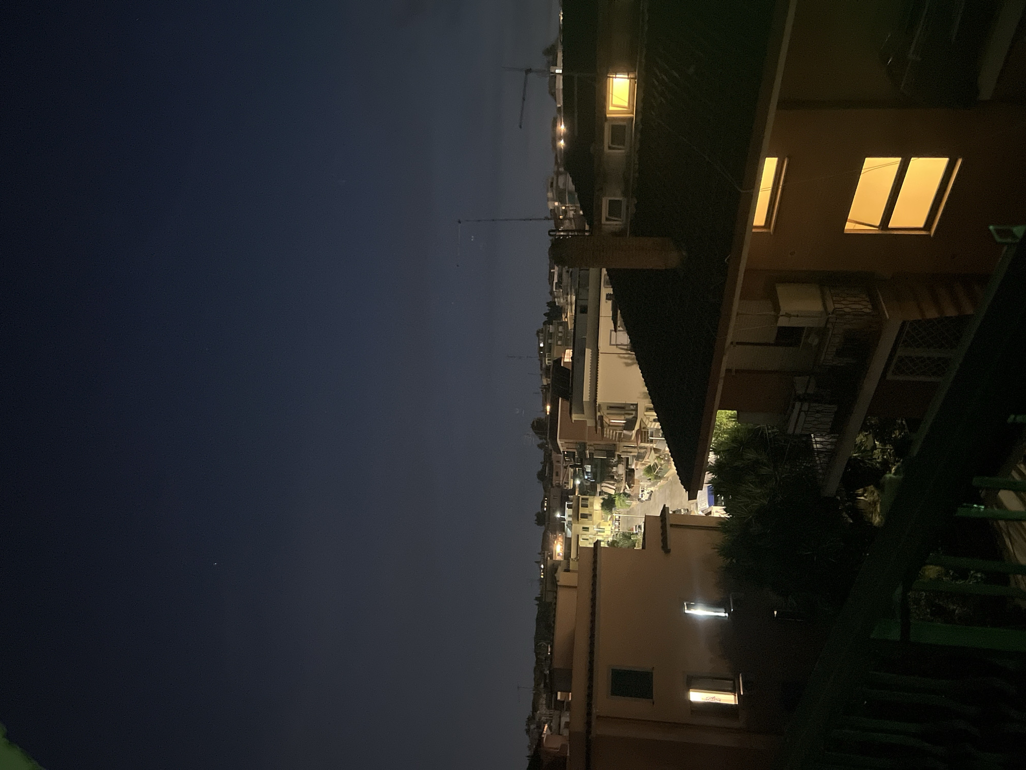 The image shows an aerial view of a residential area at night, likely in Rome, Italy, given the architectural style of the buildings and the presence of a small star in the sky. The scene is illuminated by artificial lights from within houses and streetlights along a narrow road. In the foreground, a dark green railing is visible, suggesting the vantage point is from a balcony or rooftop. Several buildings with tiled roofs are apparent, some with visible windows casting a warm yellow glow. One building in the mid-ground has an air conditioning unit mounted on its exterior. In the distance, more buildings with illuminated windows can be seen, along with what appear to be antenna towers and possibly a crane. The sky is dark blue, indicating nighttime, with a single bright star visible in the upper portion of the frame. There are no people or animals clearly discernible in the image.