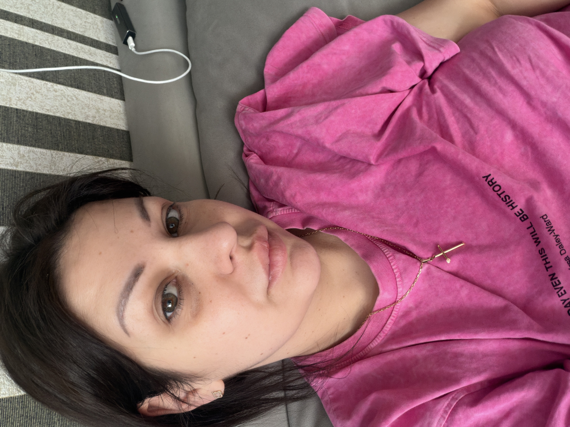 A close-up, high-angle, selfie-style shot shows a woman with dark hair and brown eyes, wearing a bright pink t-shirt with a gold cross necklace. She is lying down on a gray sofa, with a striped patterned wall in the background. A white cord and a black power bank are visible on the sofa to her right. The image provides no information about the city of Sochi, Russia.
