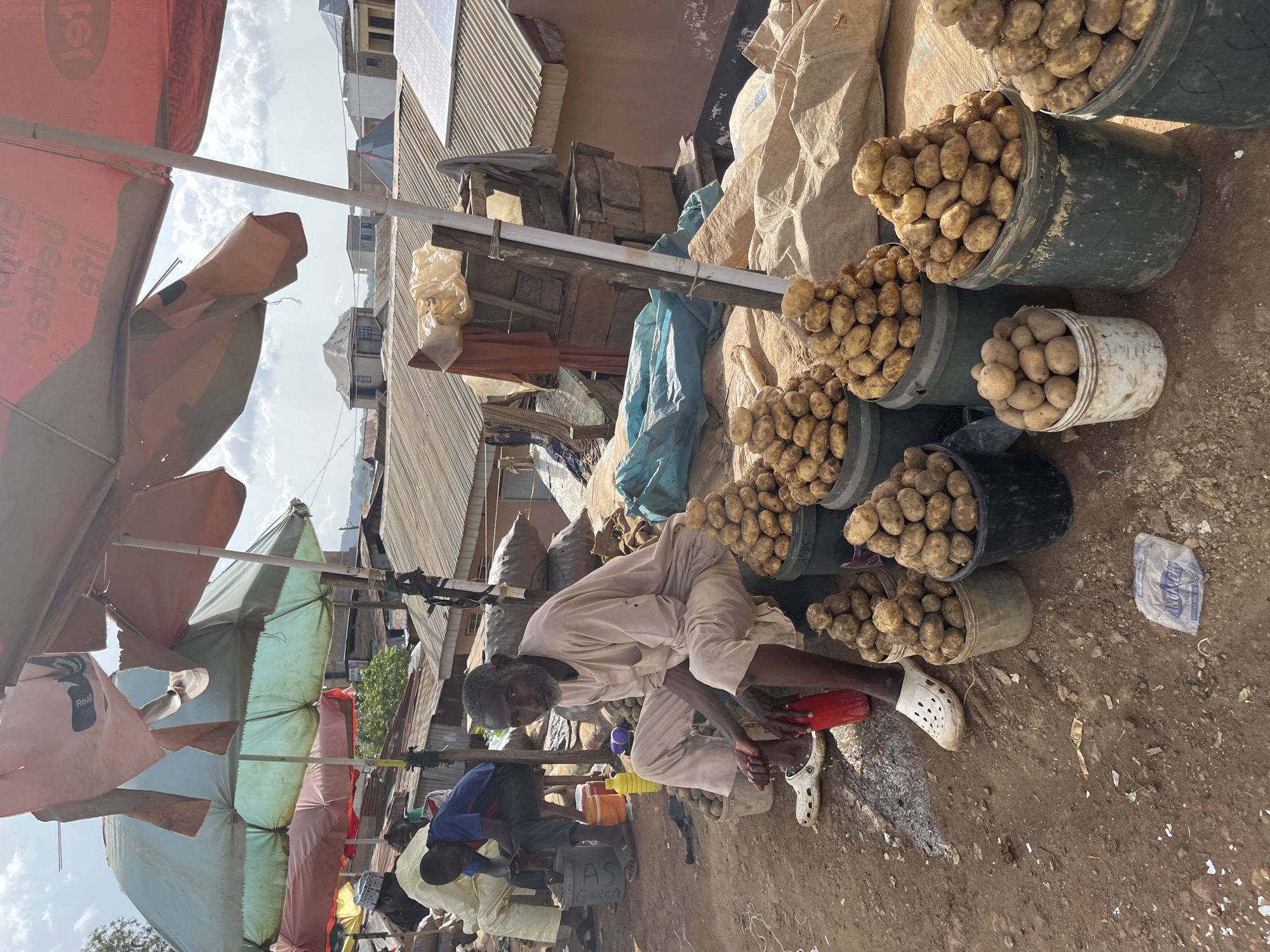 In Jos, Nigeria, an outdoor market scene unfolds under a cloudy sky. Multiple tattered umbrellas, some red and some green, provide shade. In the foreground, an elderly man with graying facial hair sits on a low stool, wearing a light-colored traditional outfit. He is tending to his feet, with one foot submerged in a red container and the other in a white clog. To his right, several buckets are overflowing with potatoes, forming neat conical piles. Behind him, another man in a blue shirt and dark trousers sits on a stool, looking down. A third man, in a light-colored robe and cap, is partially visible in the background, also seated. The ground is dirt, with scattered debris. Large sacks of produce are piled in the background, and various structures, likely market stalls and residential buildings with corrugated iron roofs, are visible in the distance, indicative of the urban setting of Jos.