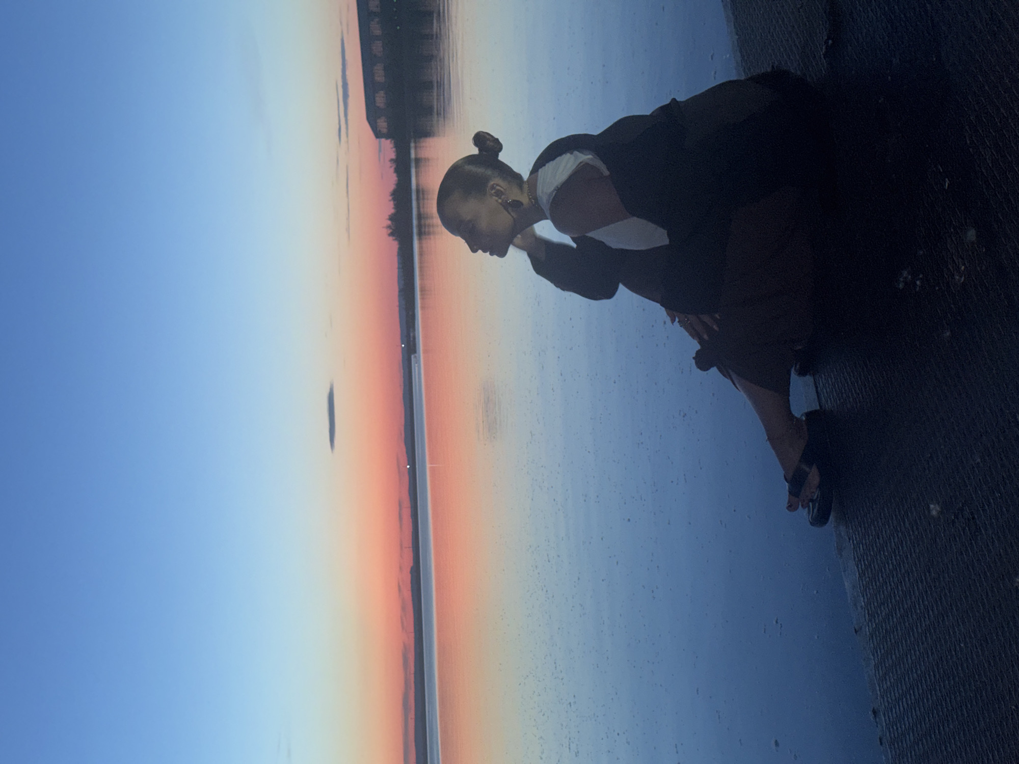 The image presents a single individual seated on a dark, textured embankment beside a large body of water at dusk in Chaikovsky, Russia. The sky transitions from a deep blue overhead to vibrant orange and pink hues along the horizon, with a dark, elongated cloud visible in the upper right. These sky colors are reflected on the subtly rippled surface of the water, which also shows numerous small, light-colored speckles. The seated individual, a female, is positioned in profile, facing right and looking upward. She has dark hair pulled into a high bun and wears an earring. Her attire consists of a white top underneath a dark open jacket, dark lower garments, and dark flat sandals. Her left hand is raised towards her neck/chin area. Across the body of water, a distant shoreline is visible, featuring silhouetted buildings, including one with multiple visible openings, and sparse tree lines, along with faint structures on the far horizon.
