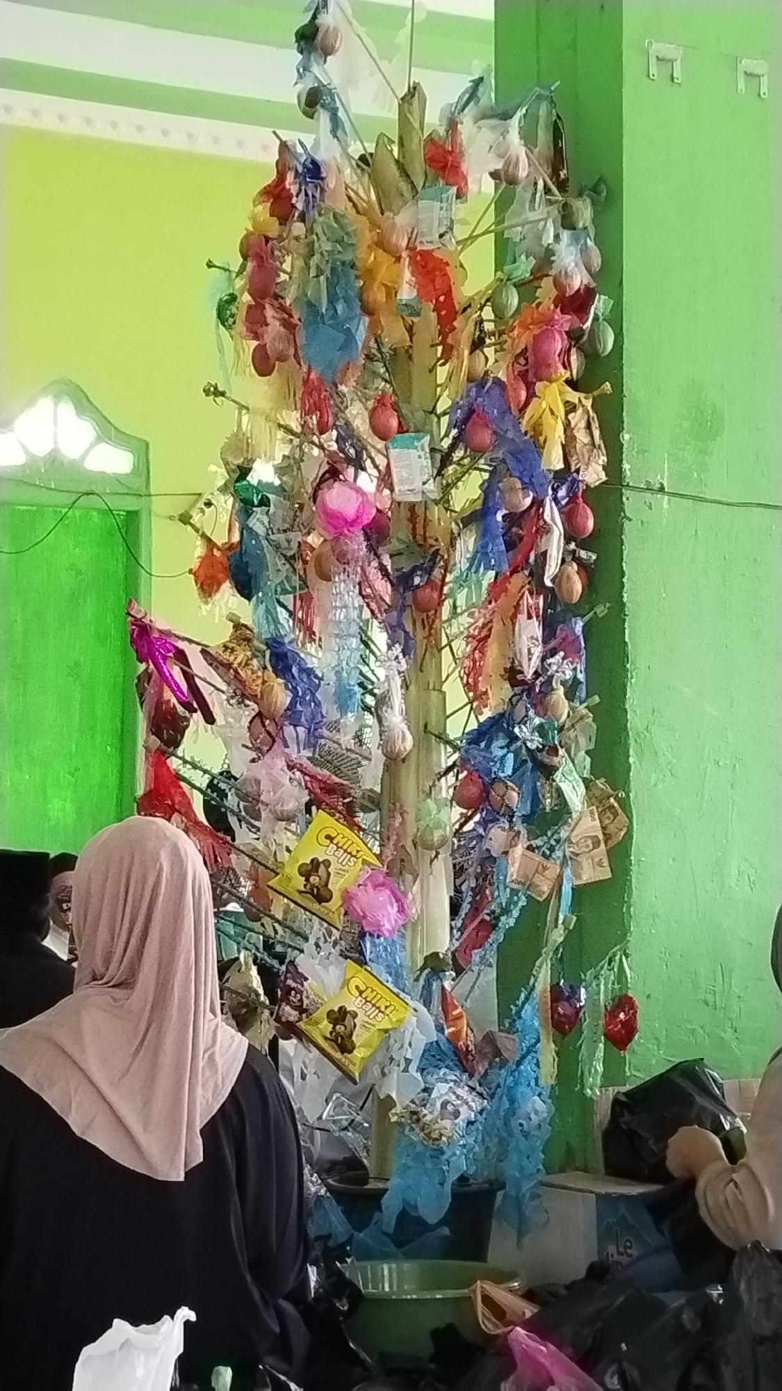 In Pangipangi, Indonesia, a brightly lit room with lime green walls serves as the backdrop for a decorated tree-like structure. The structure, made of branches or stalks, is adorned with a multitude of colorful items, including what appear to be wrapped candies, small bags, and possibly coins or money tied with string. Several individuals are present, including a woman in the foreground wearing a light pink hijab and a black garment, her back to the camera. Another person, partially visible to the left, is wearing a black head covering. In the background, other individuals can be seen, as well as various plastic bags and containers. The overall scene suggests a cultural or religious event, possibly a celebration or offering, given the decorated tree and the presence of money and food items.
