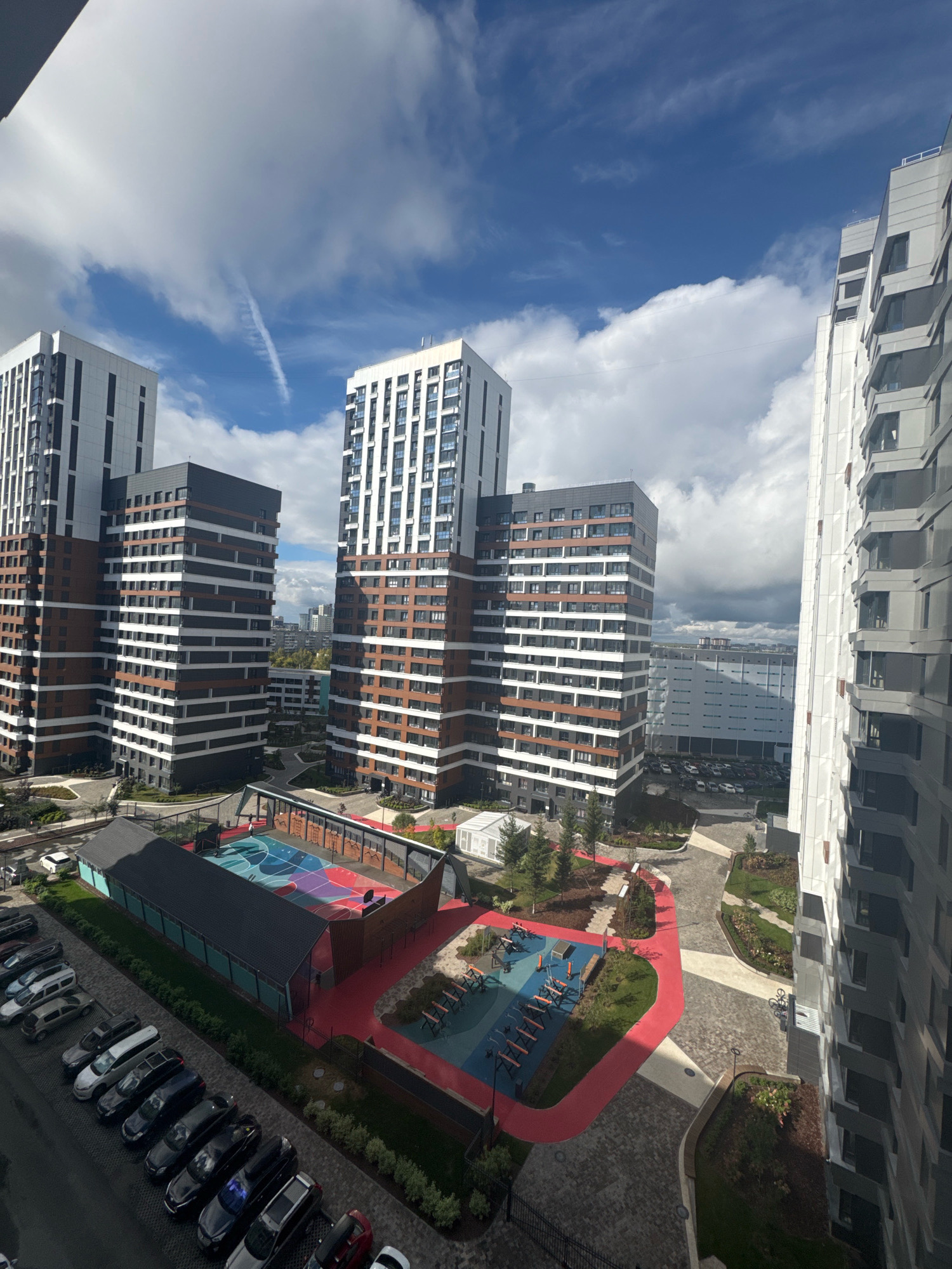 This is an aerial, high-angle, wide shot of a modern residential complex in Novosibirsk, Russia. The scene features several tall, multi-story apartment buildings with a distinctive facade of alternating brown and white horizontal bands. In the foreground, there is a sports court with colorful markings, a basketball hoop, and surrounding benches and playground equipment. Adjacent to the court is a long, dark-colored structure with a pitched roof. A line of parked cars is visible on a paved area to the left. The ground is a mosaic of different paving stones and landscaped green spaces with trees. The sky above is partly cloudy with streaks of contrails from an aircraft. The overall impression is one of a newly developed urban residential area.