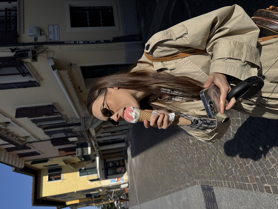 A female subject is depicted mid-action, licking a two-scoop ice cream cone (one pink/red scoop, one white/cream scoop) on Via Don Lenotti, Peschiera del Garda, Italy. The subject, wearing sunglasses, a plaid shirt, and a beige trench coat, holds the ice cream cone in her left hand. Her right hand simultaneously grips a silver-toned smartphone in a clear case and a black retractable leash. A brown shoulder bag with a distinct pattern is partially visible on her right side. The scene is set on a cobblestone street under strong sunlight, casting prominent shadows. Background elements include multi-story buildings: a yellow-facade building with a black wrought-iron balcony on the left, and a beige-facade building with brown window shutters and a white awning on the right. The street sign "VIA DON LENOTTI" is affixed to the beige building. A clear blue sky is visible at the top of the frame.