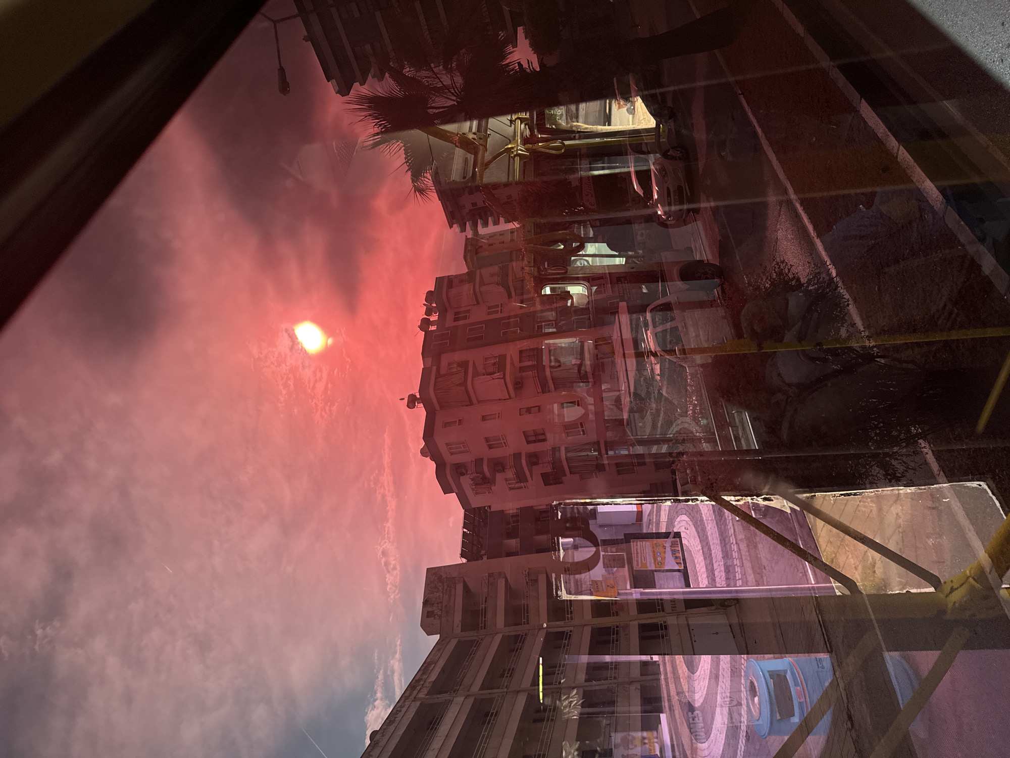 This image captures an urban street scene in Alanya, Turkey, viewed through a vehicle window, resulting in significant overlaying reflections. The sky is characterized by an intense red and pink hue, indicative of sunset or sunrise conditions, with a bright, diffuse light source, likely the sun, in the upper-center, and scattered clouds containing two visible contrails.
The foreground is dominated by strong reflections of the interior of the viewing vehicle, specifically showing yellow metallic vertical and horizontal support bars and overhead handrails, which obscure parts of the external environment. Indistinct, faint reflections of individuals or objects within the vehicle are also present.
The midground details a street with an asphalt surface and a partial white lane marking. A white pickup truck and a light-colored sedan (resembling a Hyundai i20) are parked along the right side of the street. The sidewalk features a distinctive circular paving pattern and a blue cylindrical public trash bin. Multiple multi-story buildings line the street; the structure on the left exhibits numerous balconies and appears somewhat weathered, while a building on the right also features balconies and rooftop satellite dishes. Several palm trees and other sparse foliage are interspersed within the urban setting. A street light fixture is visible extending from a building on the right. Partial commercial signage with Turkish text, such as "SİXAT" and "TELER", is discernible on building facades. No specific human actions or interactions are clearly depicted, beyond the implied presence of occupants within the viewing vehicle.