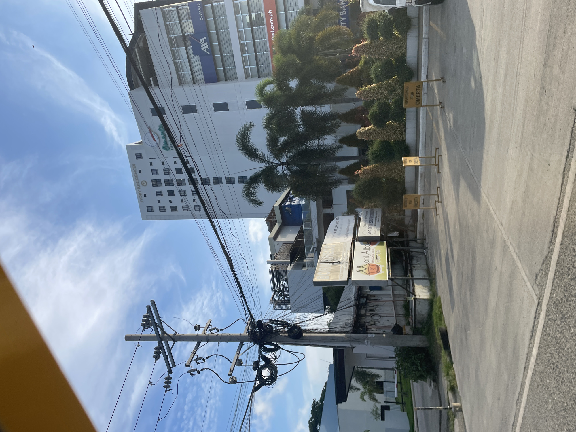 In Davao City, Philippines, a street scene unfolds under a bright, partly cloudy sky. A prominent utility pole, laden with a complex network of electrical wires, stands near the foreground on the left. Behind it, a modern, multi-story white building, labeled "IVORY RESIDENCES," dominates the skyline. The building features numerous dark-paned windows and a circular green logo with the text "Elements." To its right, another section of the building displays blue signage with "AXA" and "DAVAO," and orange signage that reads "fwd.com.ph." Several palm trees and manicured shrubs are visible in front of the buildings. In the middle ground, several yellow signs with black text, reading "RESERVED FOR OMERTA," are placed on stands, indicating designated parking spots. The ground in the foreground and middle ground is paved with concrete.
