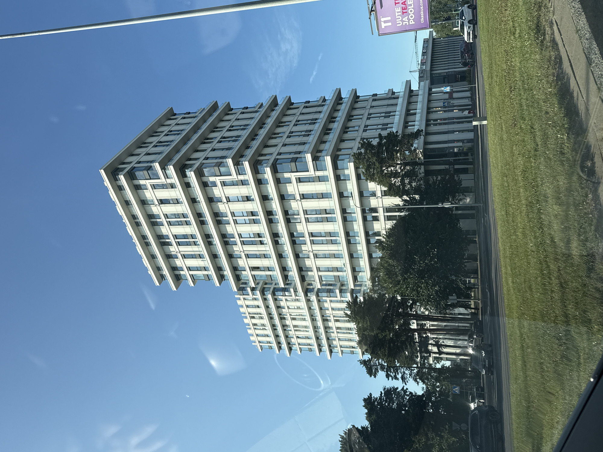 The image shows a modern, multi-story building in Tallinn, Estonia, with a clear blue sky in the background. The building has a white facade with many windows and balconies. Trees are visible in front of the building, and a road with cars is in the foreground. A tall light pole is also visible to the right of the building. A billboard with the text "TI" and some Estonian text is also present.